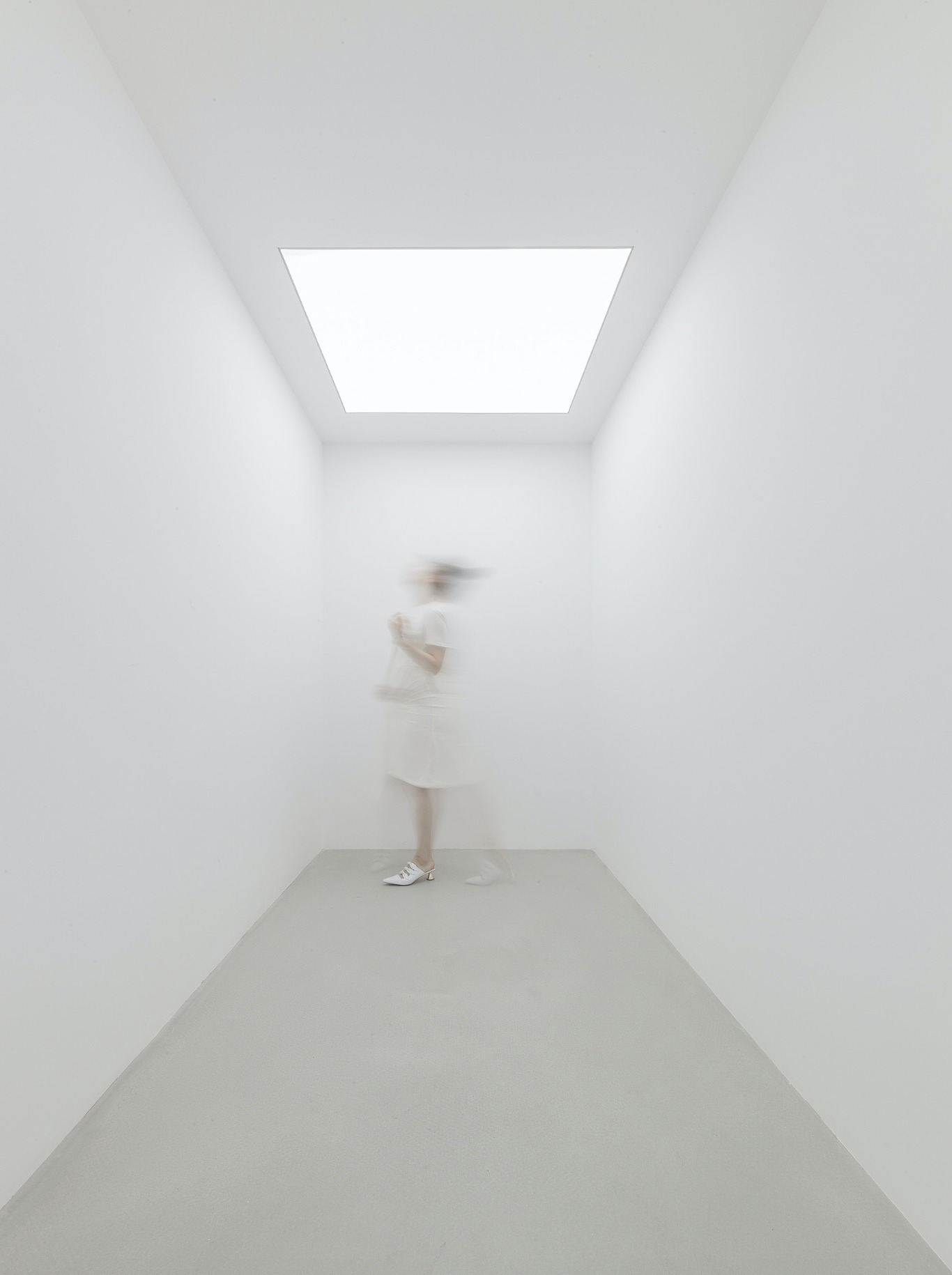 Narrow white room with a square skylight-like ceiling panel and a blurred figure in white standing against the back wall