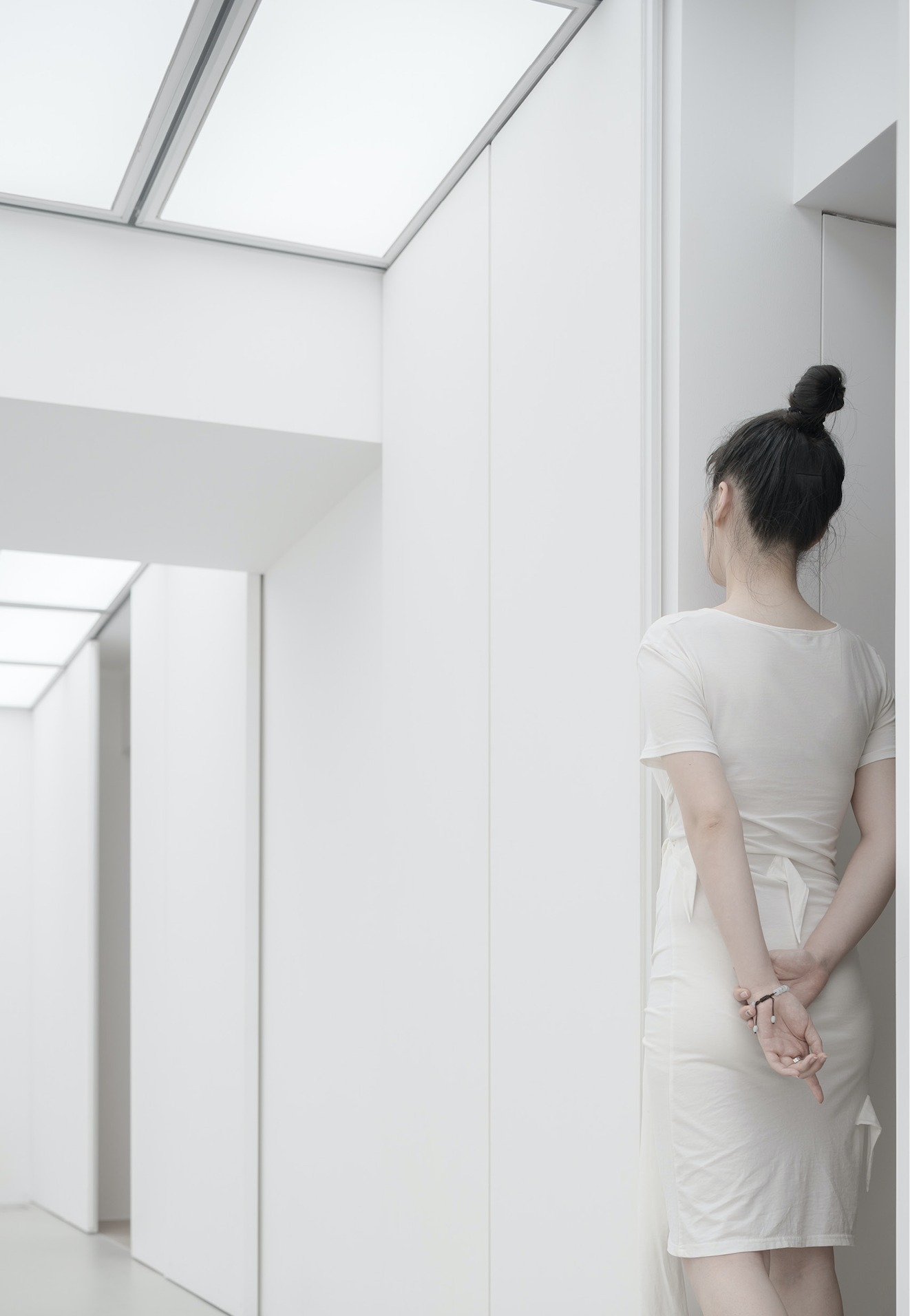 Woman in a white dress standing with hands behind her back in a white corridor under glowing ceiling light panels