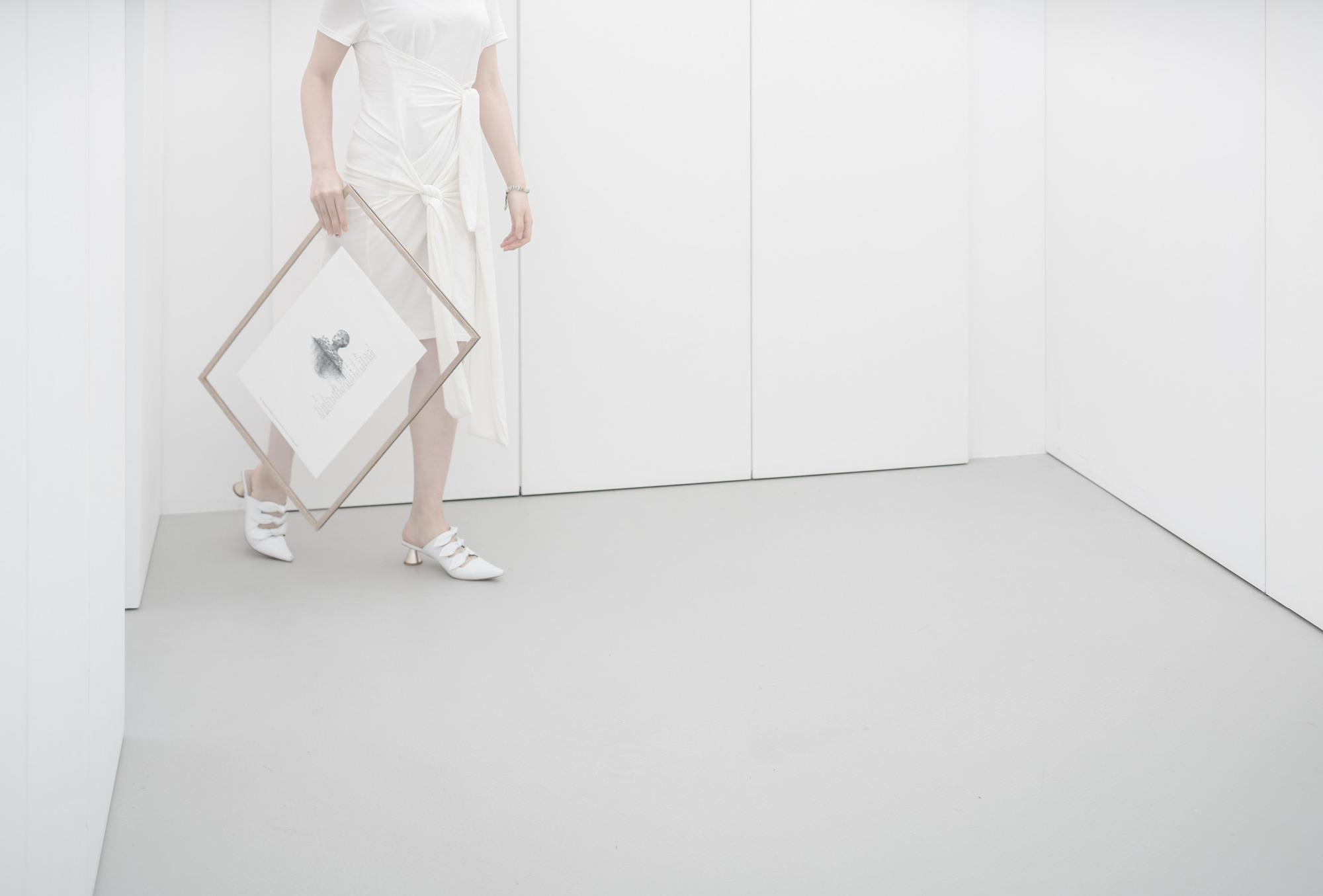Woman in white carrying a diamond-oriented framed artwork across a grey floor in an empty white gallery room