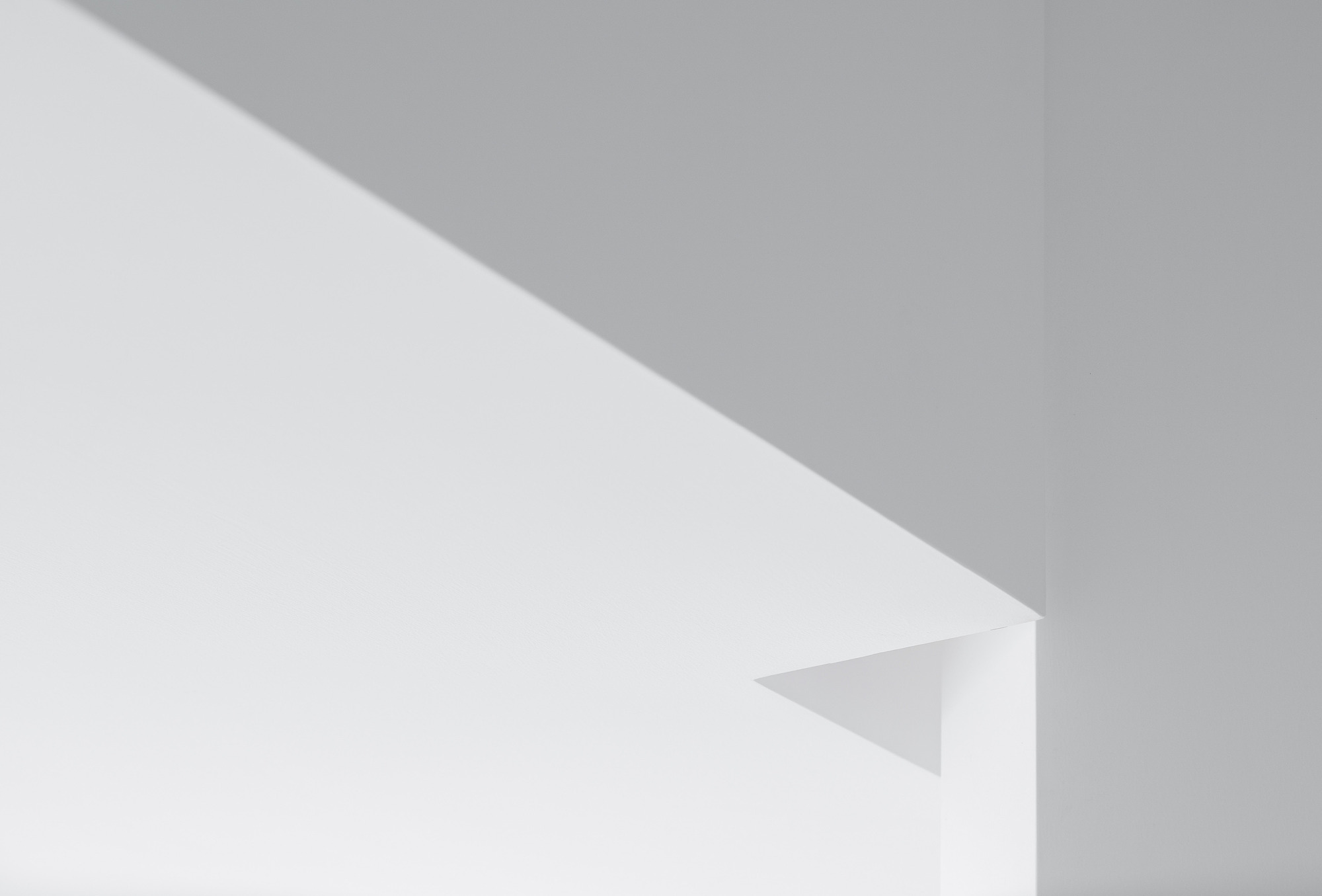 Abstract close-up of a white wall corner with a small triangular notch, split by a diagonal shadow in soft grey light