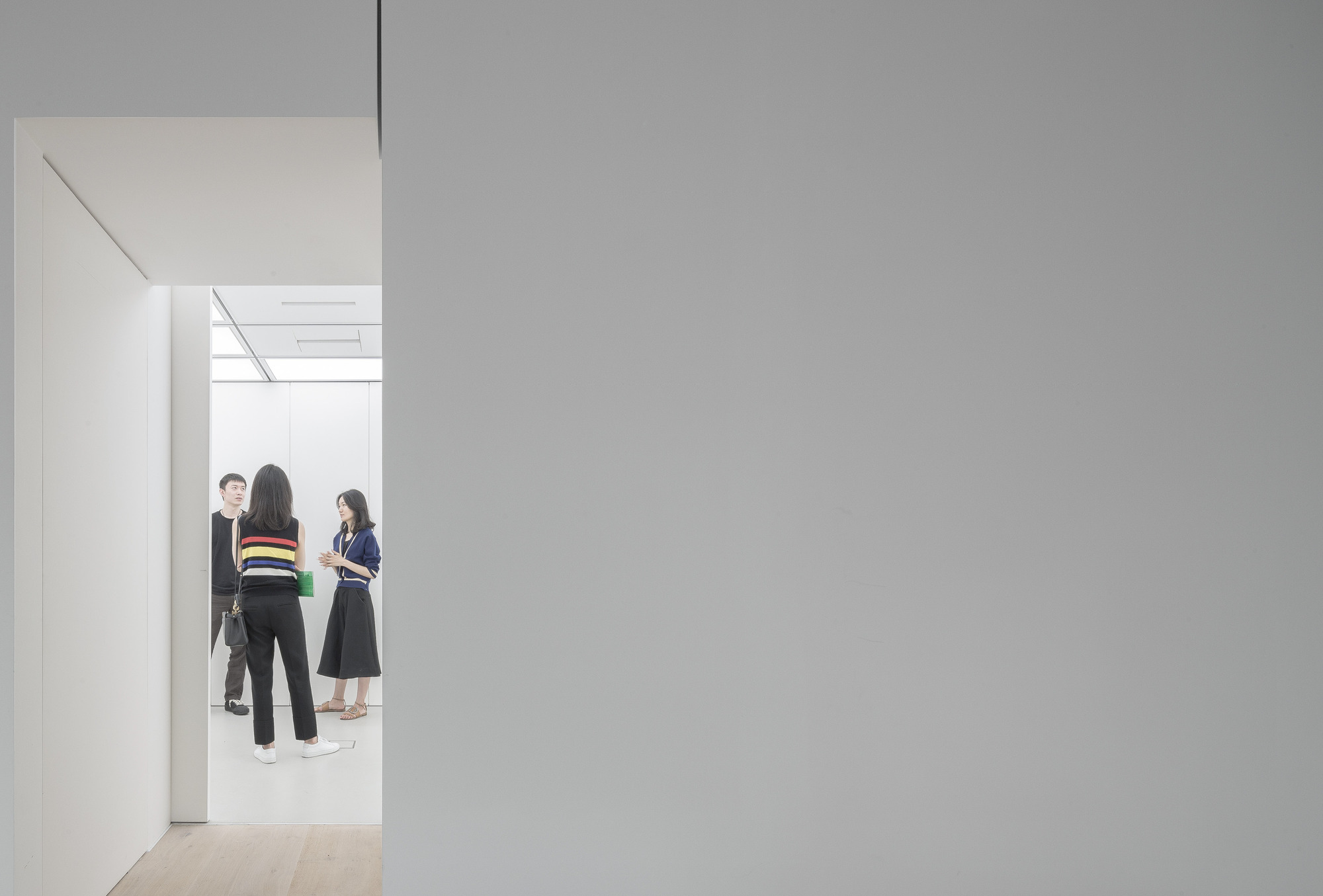 Three visitors chatting in a bright gallery room, seen through a doorway cut in a grey wall beside a pale timber floor