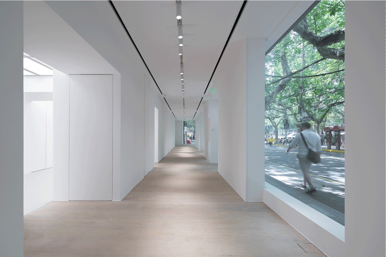 Same white corridor with a pedestrian carrying a shoulder bag passing the tall street window beneath leafy branches