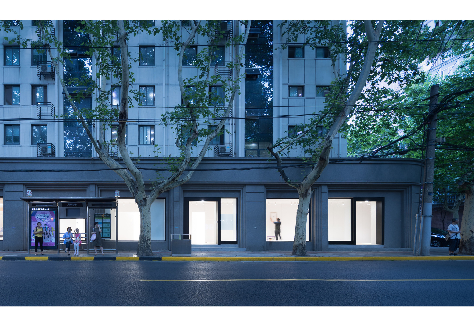 Street facade of a grey apartment block's ground floor with lit gallery windows, plane trees and a bus shelter at dusk