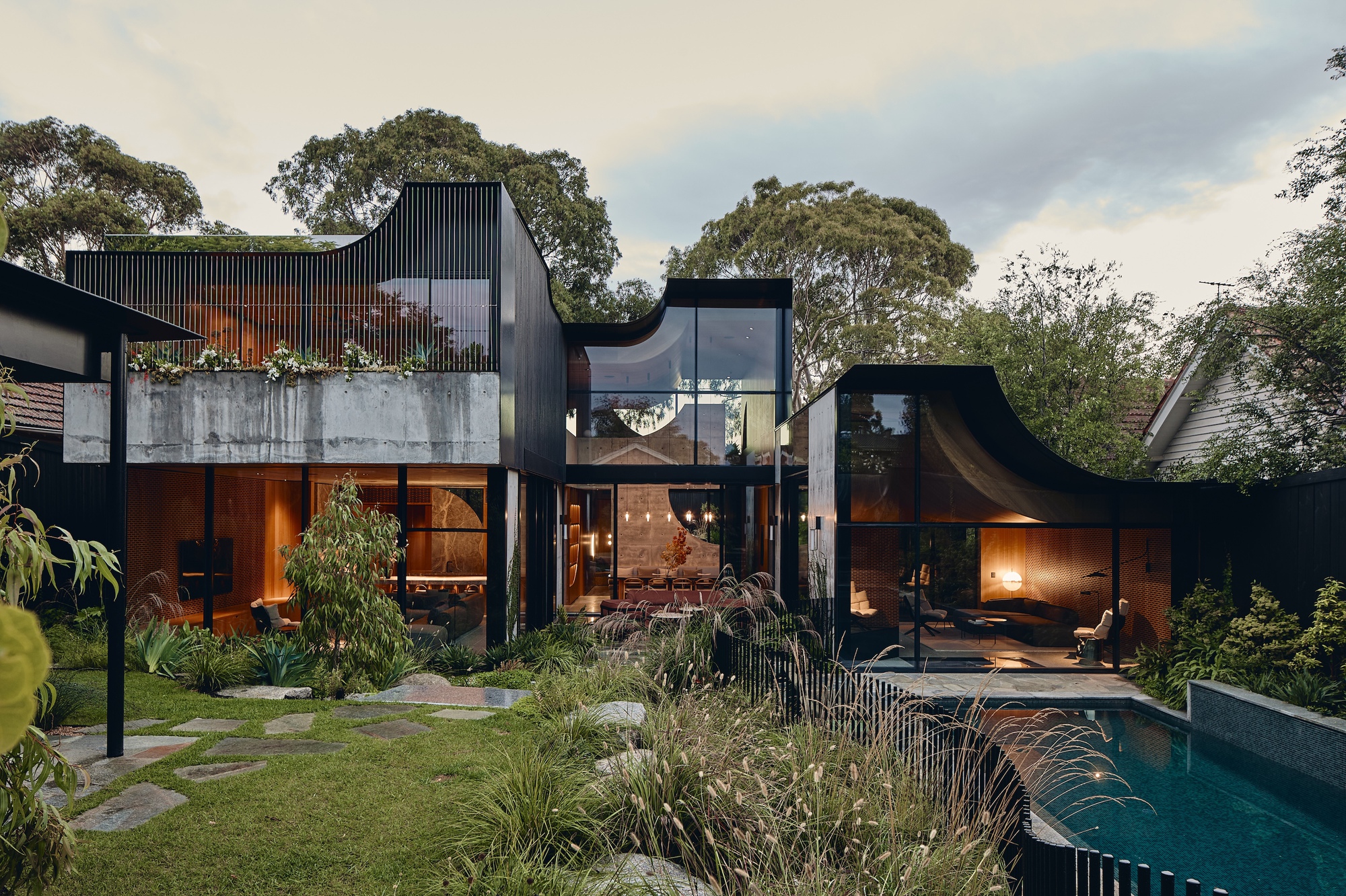 Garden facade of dark timber-clad volumes with curved rooflines above a concrete planter, stepping stones and pool at dusk