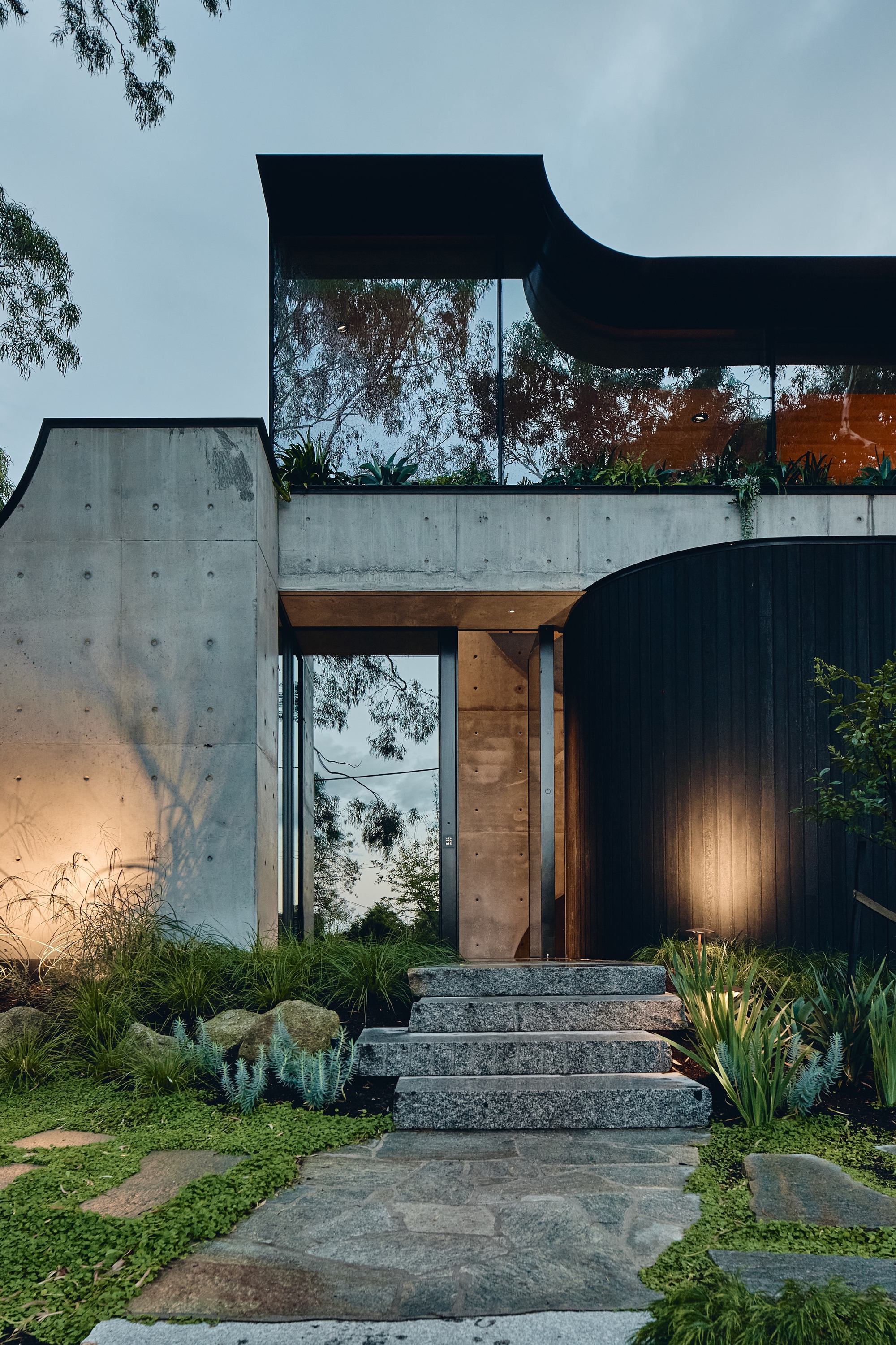 Granite entry steps rise between a board-marked concrete wall and a curved black timber wall, lit by low garden lights at dusk
