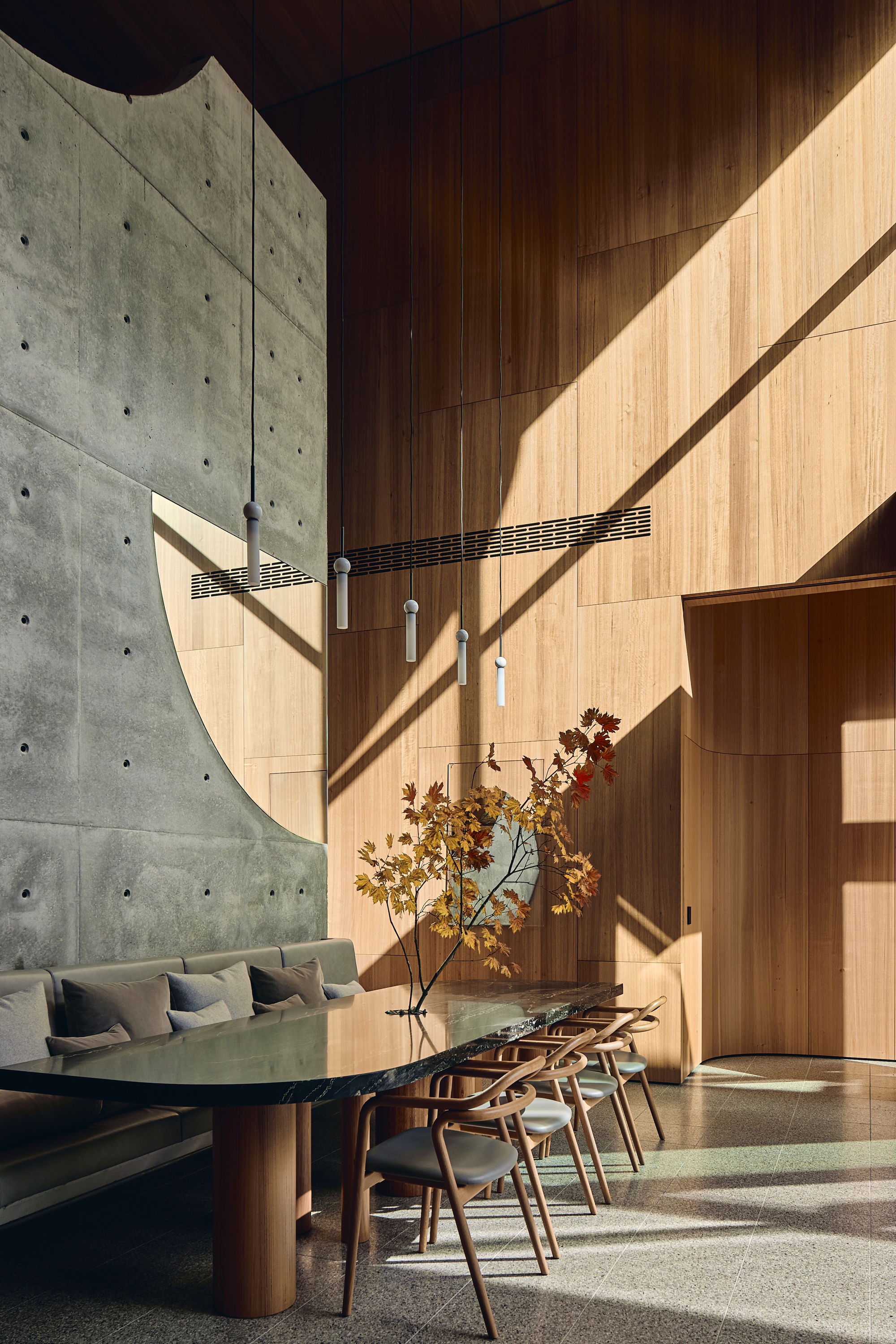 Dining table and curved banquette beneath a tall timber-panelled wall and cast concrete, with sun streaking across the surfaces