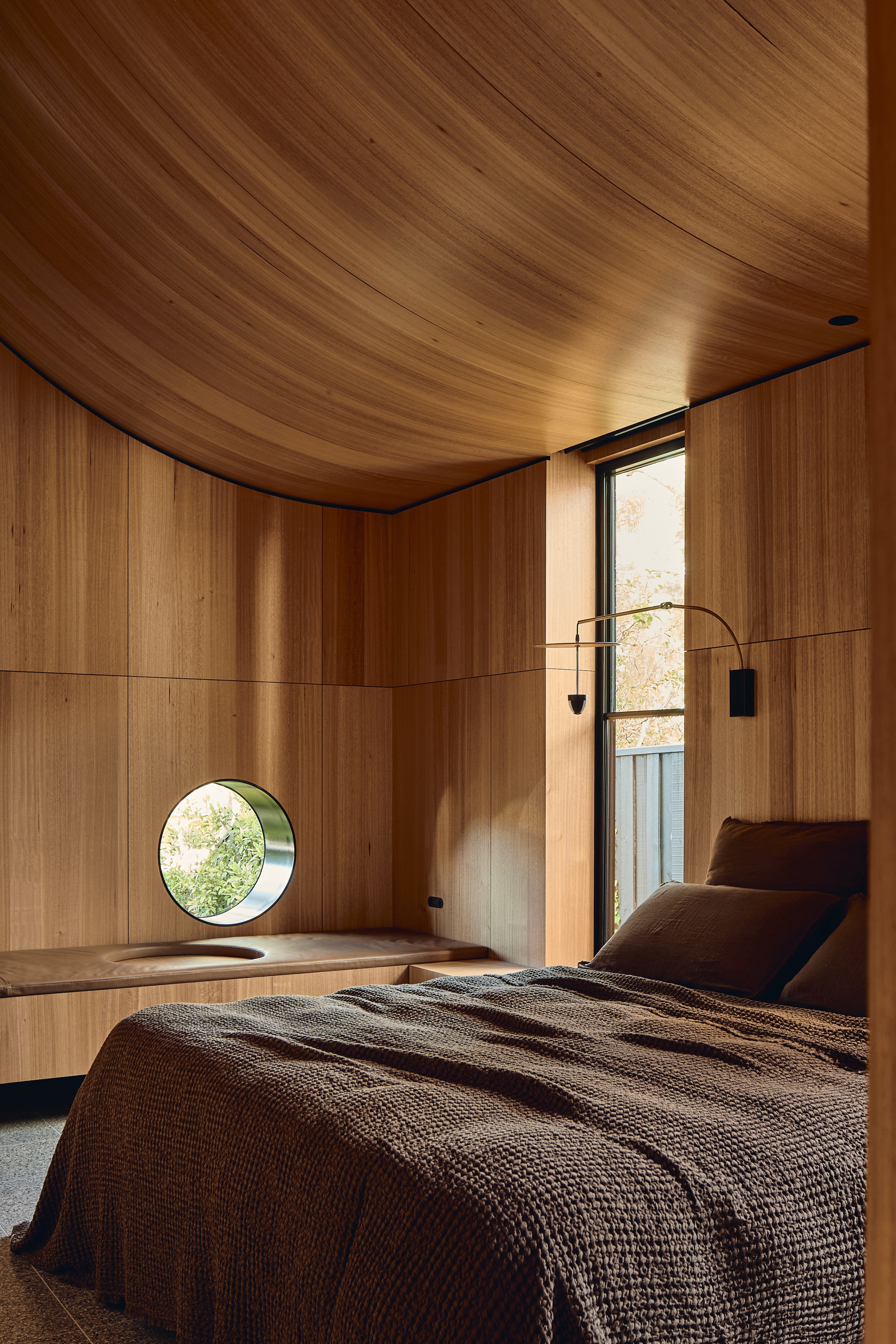 Bedroom lined in timber with a curved ceiling, round window and built-in window seat beside a tall glazed door