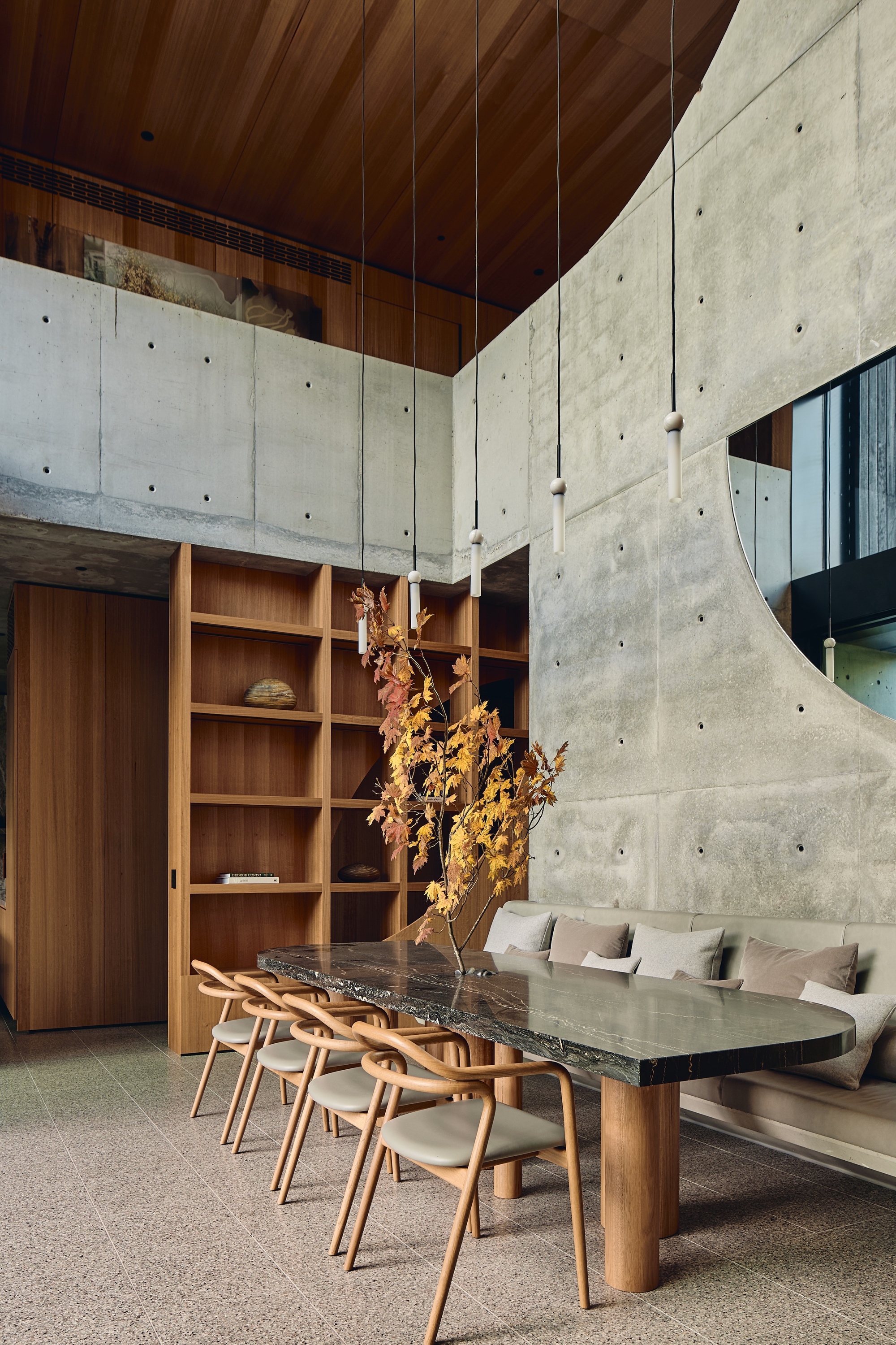 Double-height dining area with board-marked concrete walls, timber shelving and a stone-topped table holding a branch of autumn leaves