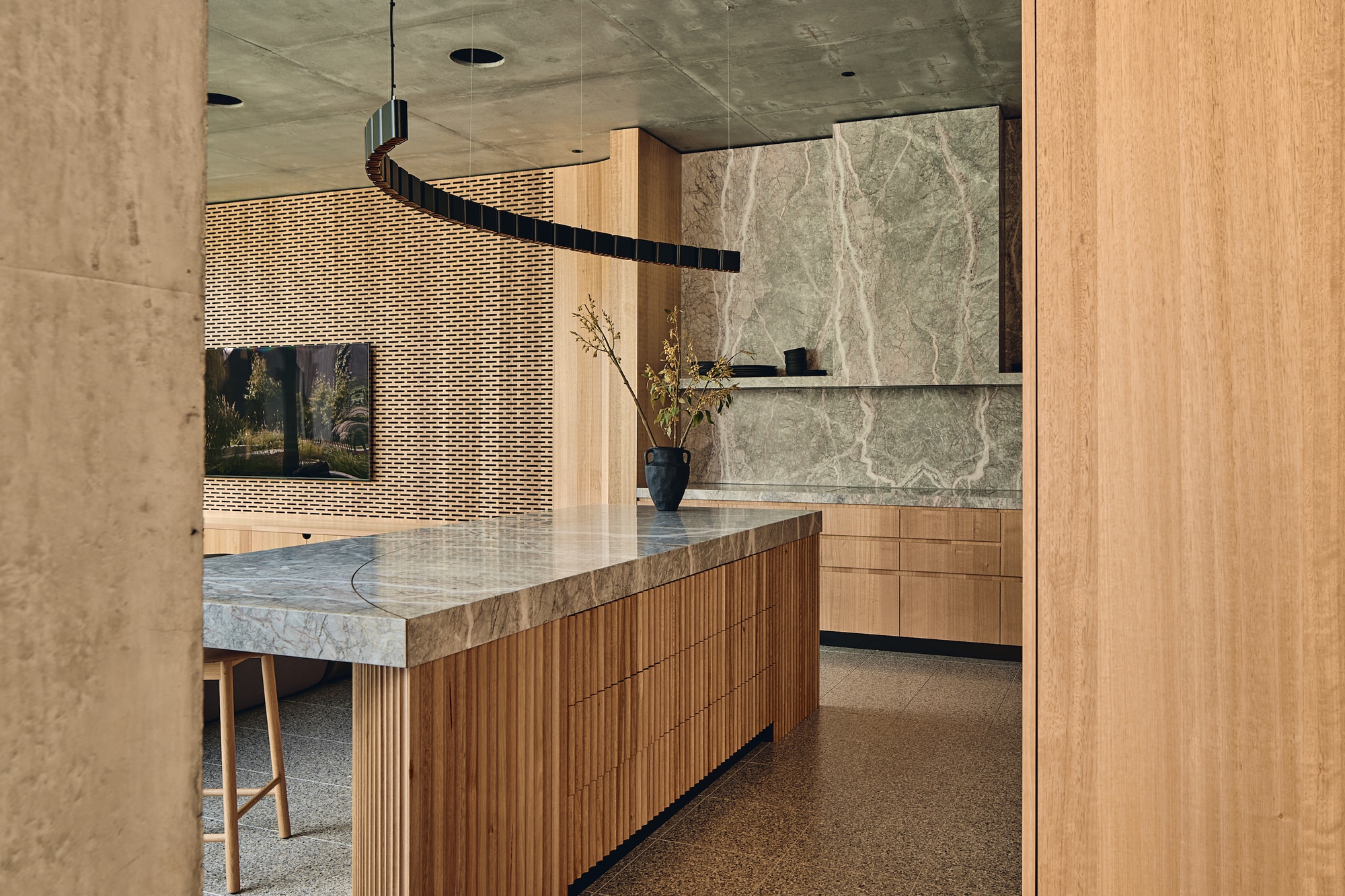 Kitchen island clad in vertical timber fluting with a stone top, set before a veined green stone splashback and perforated brick wall