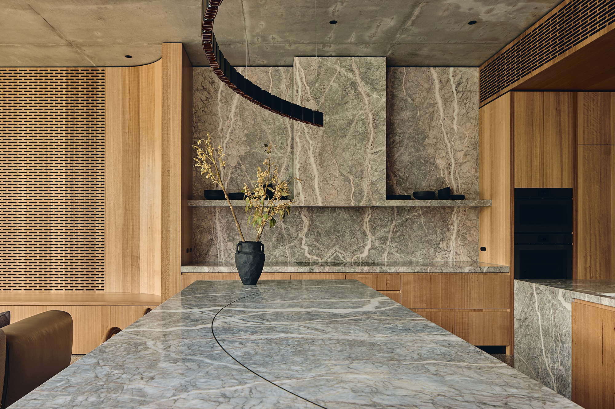 Veined green stone island top and splashback beside curved timber joinery, with a dark vase of dried branches in soft light
