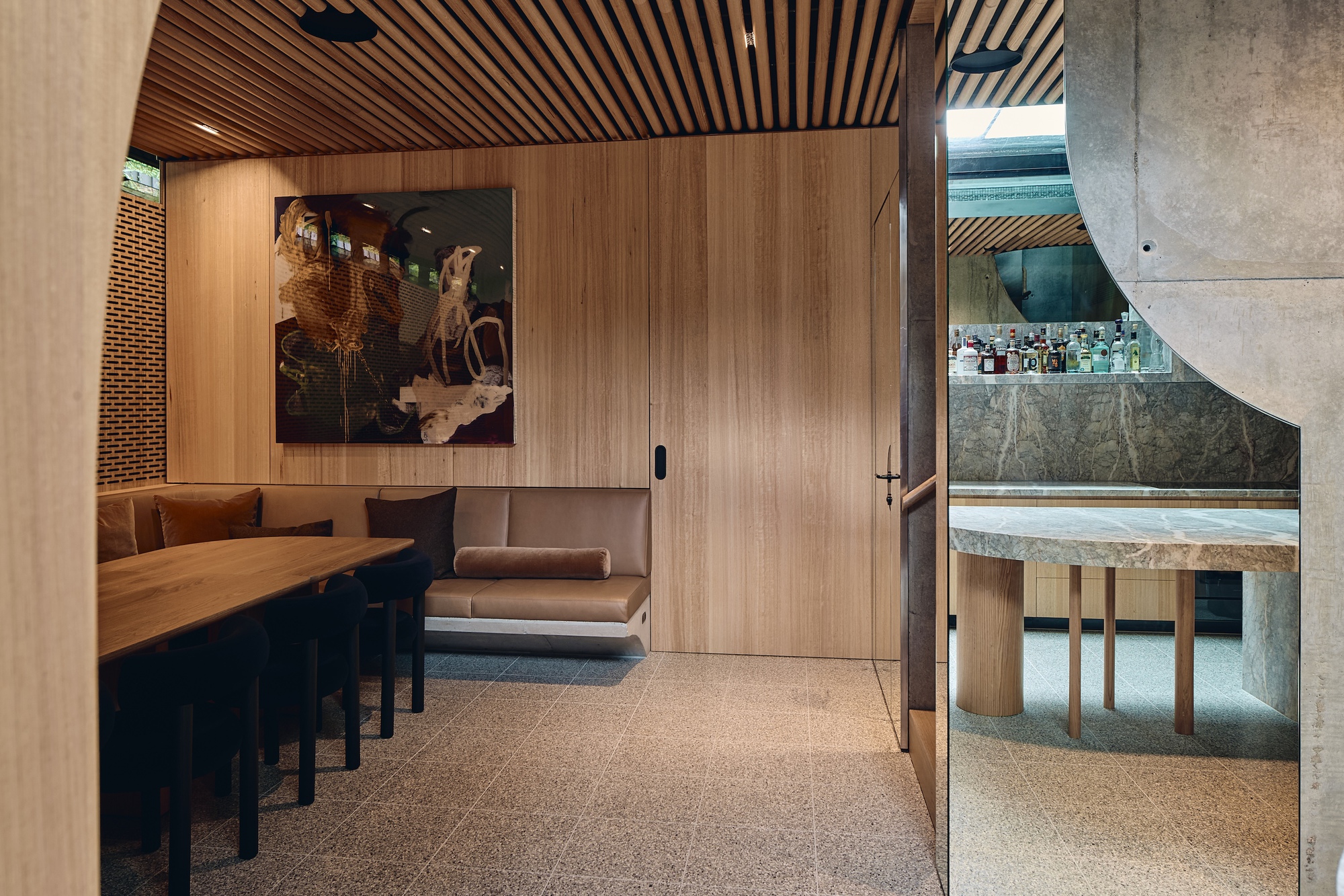 Timber-panelled dining nook with a built-in leather banquette and large painting, and a mirrored bar reflected beside a concrete arch