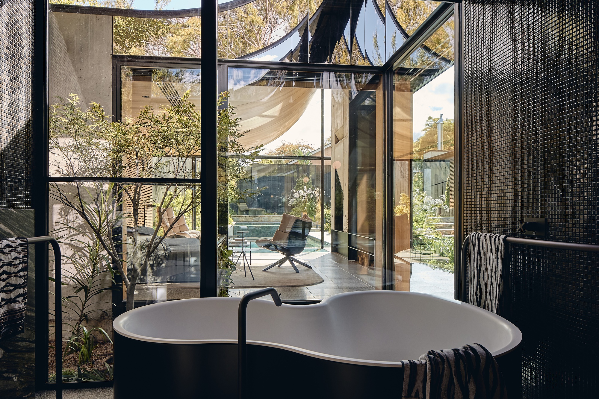 Freestanding oval bathtub framed by black mosaic tile walls and steel-framed glazing, opening onto a sunlit courtyard with a pool