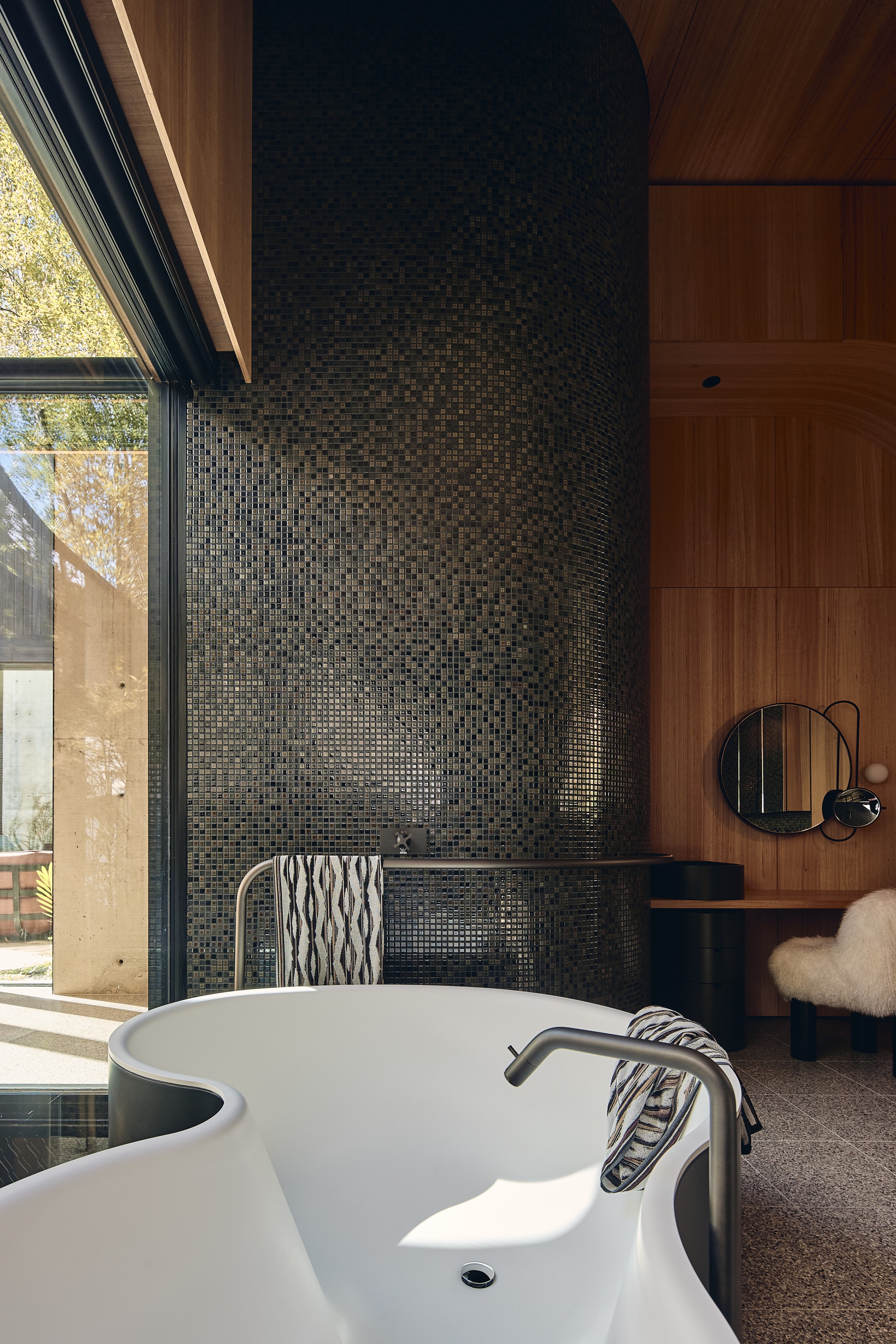 Curved black mosaic tile wall rising behind a white bathtub, with timber panelling and a round mirror in afternoon sun