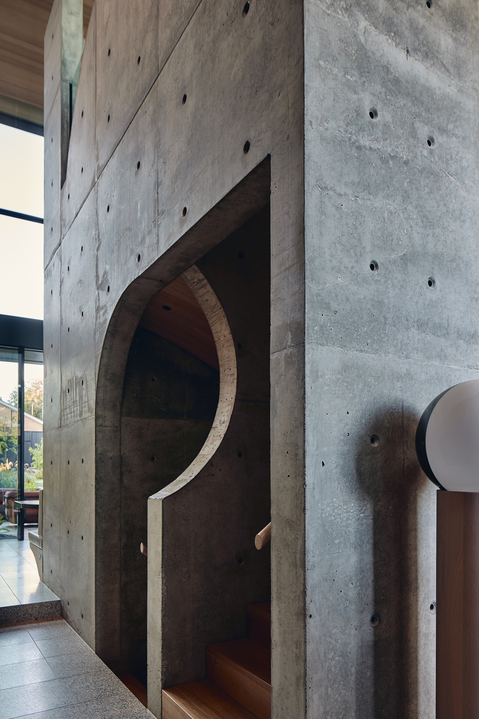 Board-marked concrete stair volume with an arched cutout, timber treads and a handrail beside a terrazzo floor