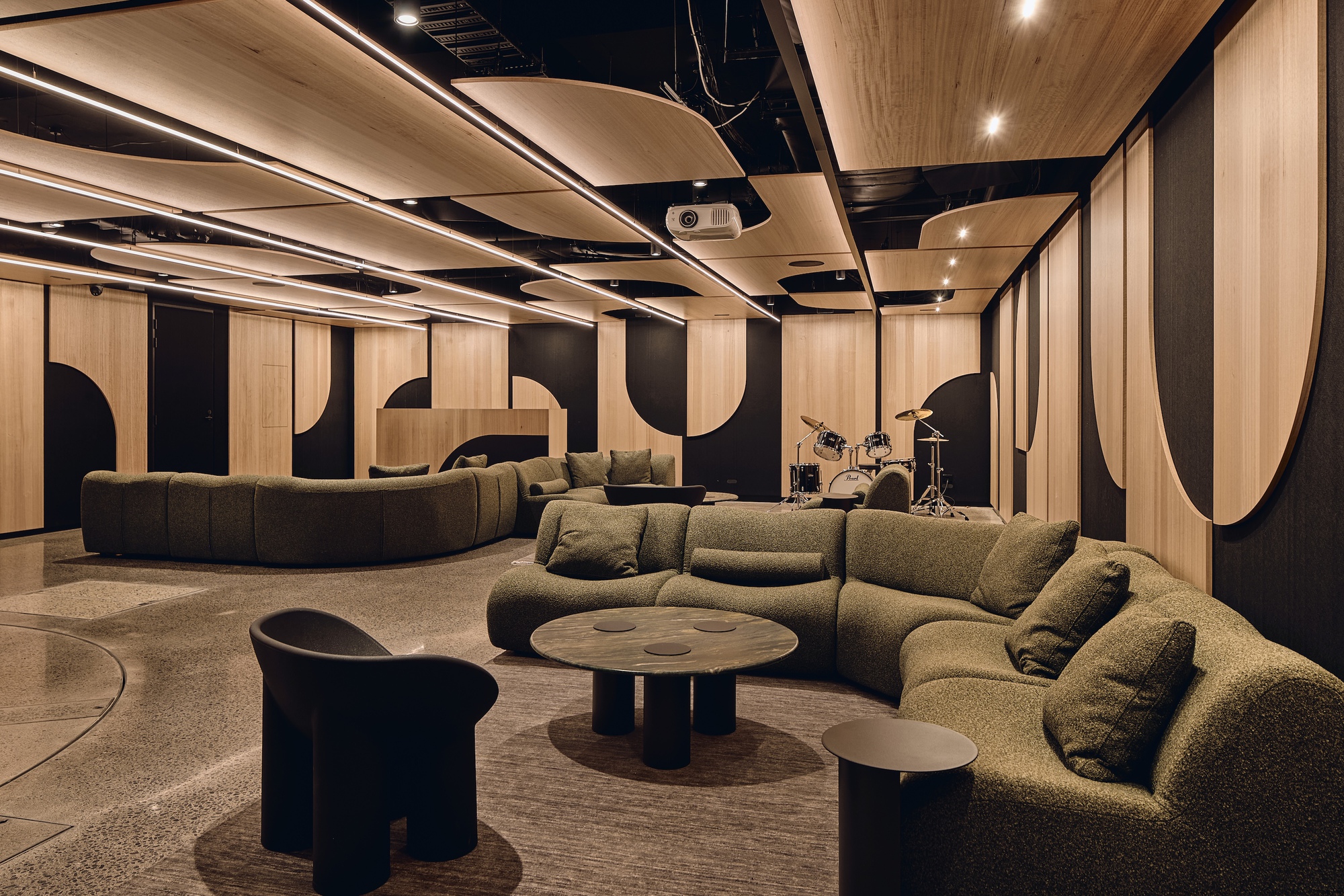 Lounge with olive curved sofas, timber and black wall panels, a drum kit and floating oval ceiling baffles