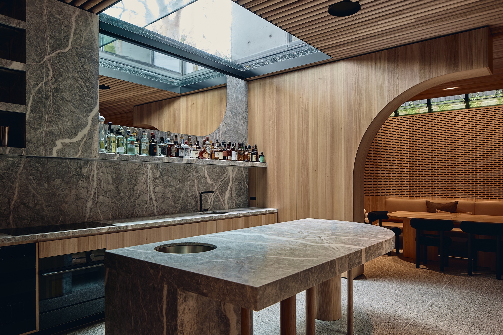 Marble-topped island and bar with a mirrored bottle shelf, timber arch and a banquette beyond, lit by a skylight