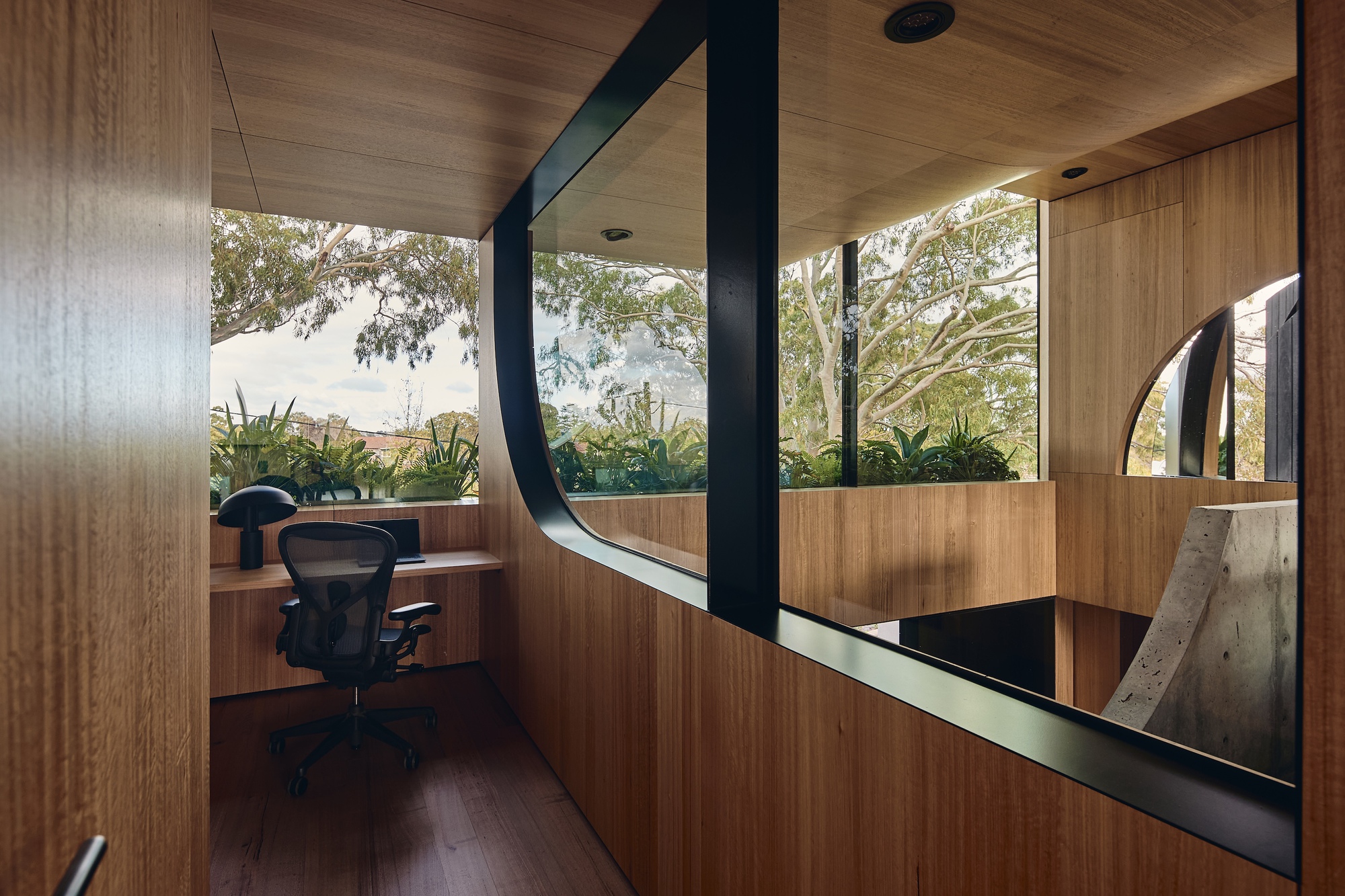 Timber-lined study nook with a desk and mesh chair, a curved black window frame looking into eucalyptus branches