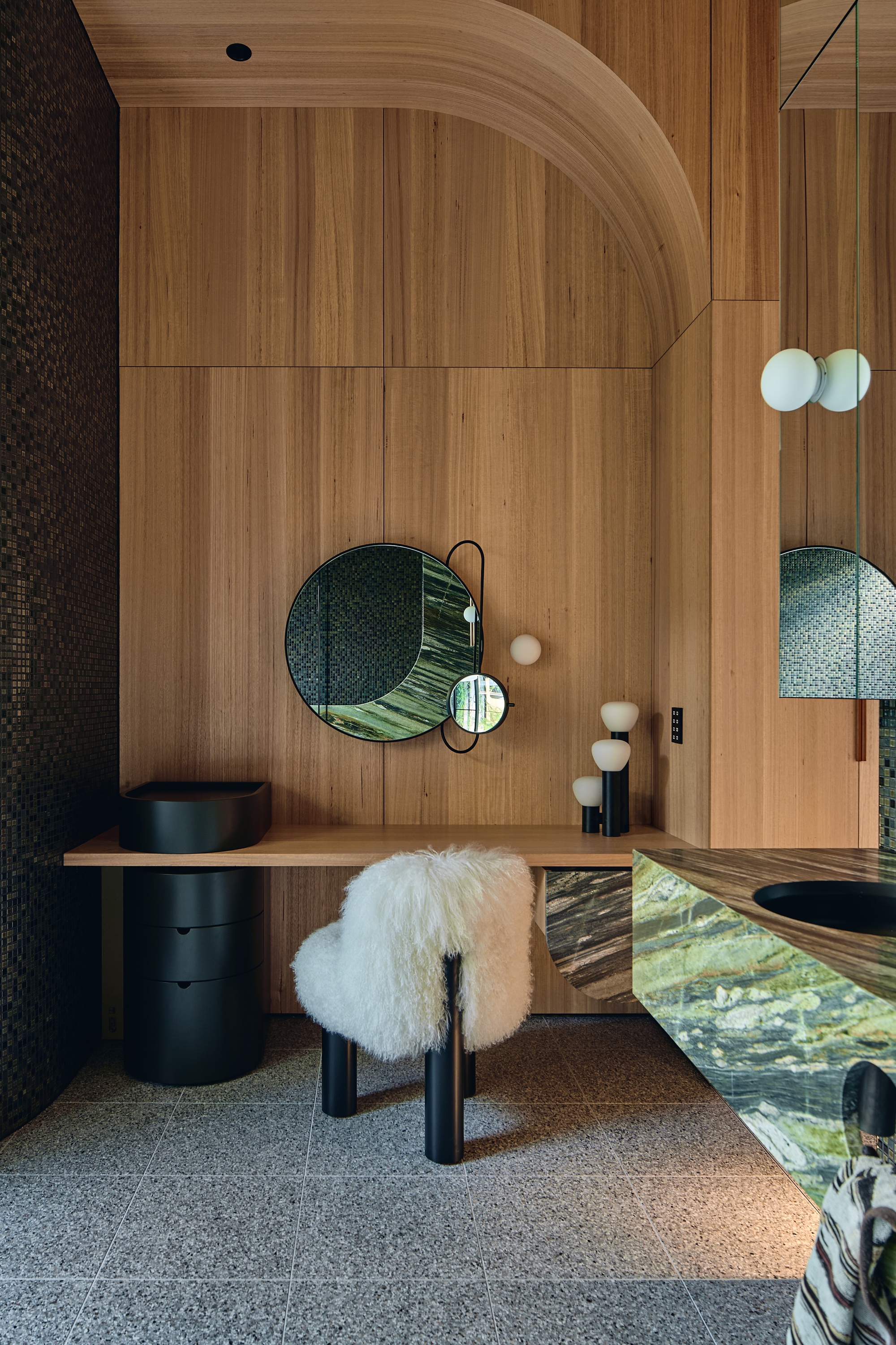 Timber-panelled dressing nook with an arched ceiling, round mirror, shaggy white stool and green marble vanity edge beside dark mosaic tile