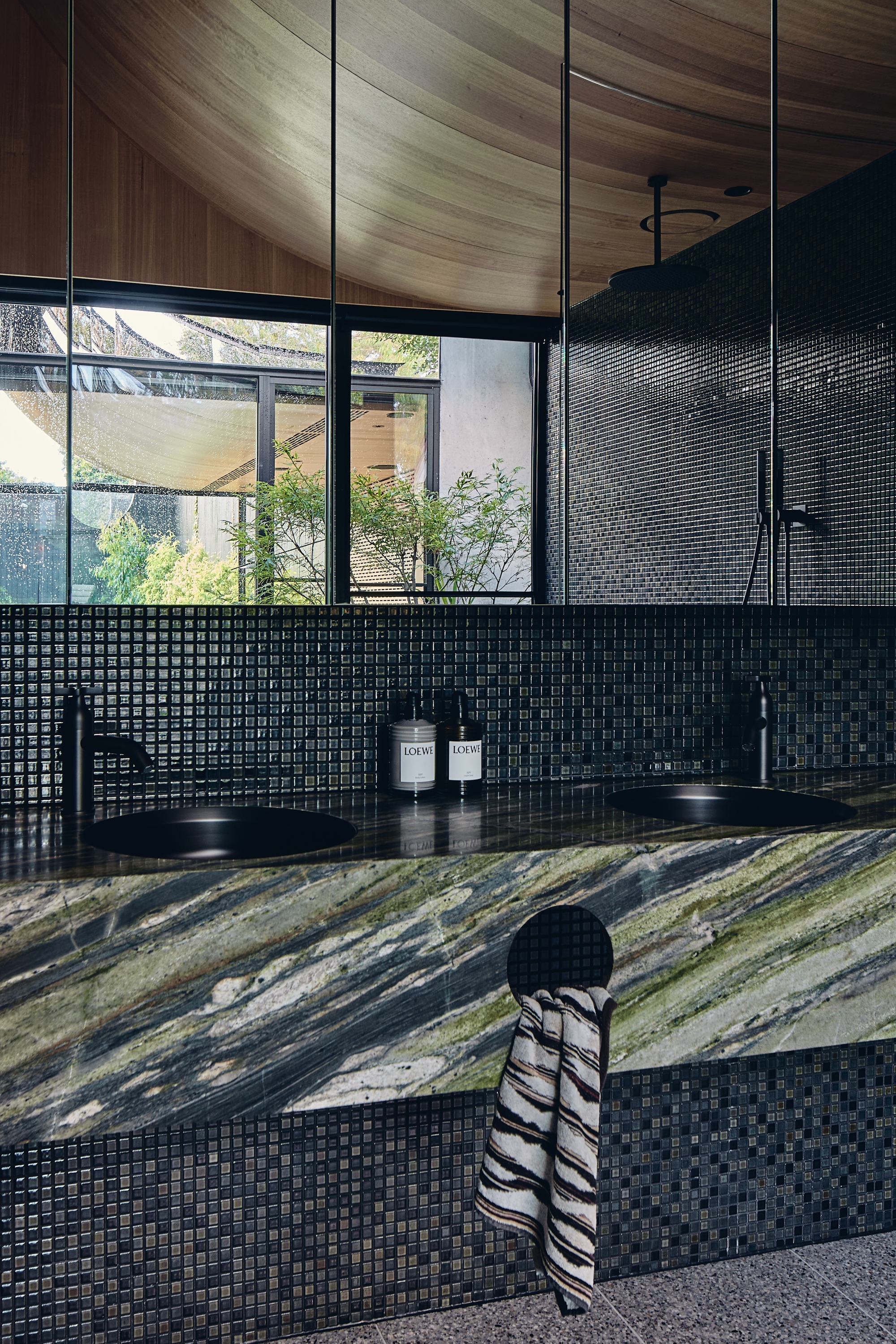 Twin black basins set in a green veined marble counter with dark mosaic tile, towel and garden glimpsed through the mirror