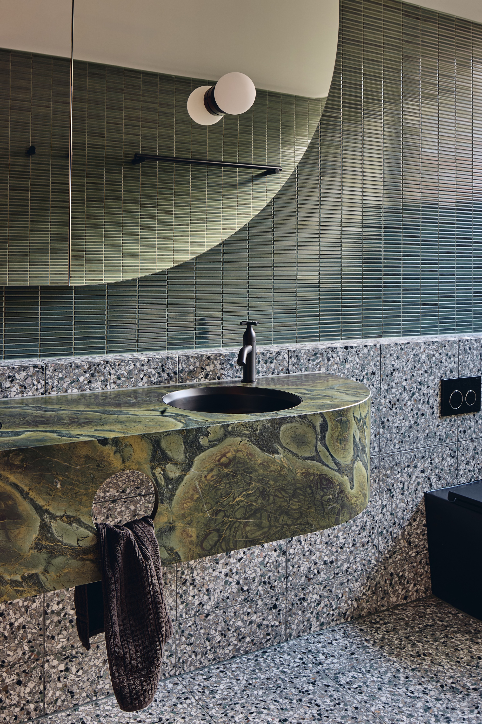Curved green marble wall-hung basin against terrazzo tile and green vertical tile, lit by a globe lamp above the mirror