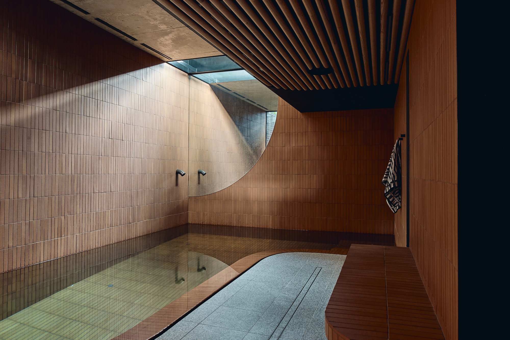 Sunken bathing pool lined with terracotta tile, a curved wall, slatted timber ceiling and a skylight casting daylight across the room