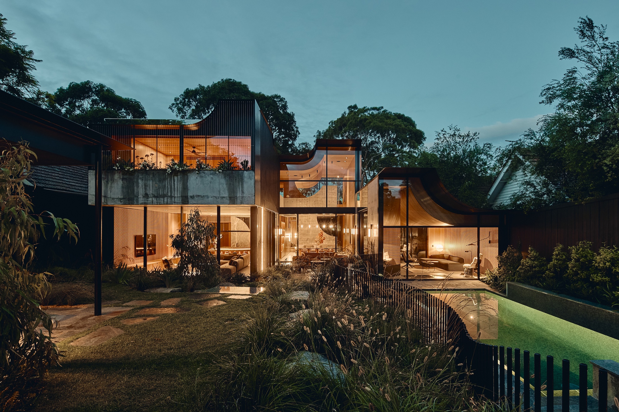 Rear facade at dusk with glowing glazed living rooms, curved roof edges, a lawn with grasses and a lit pool