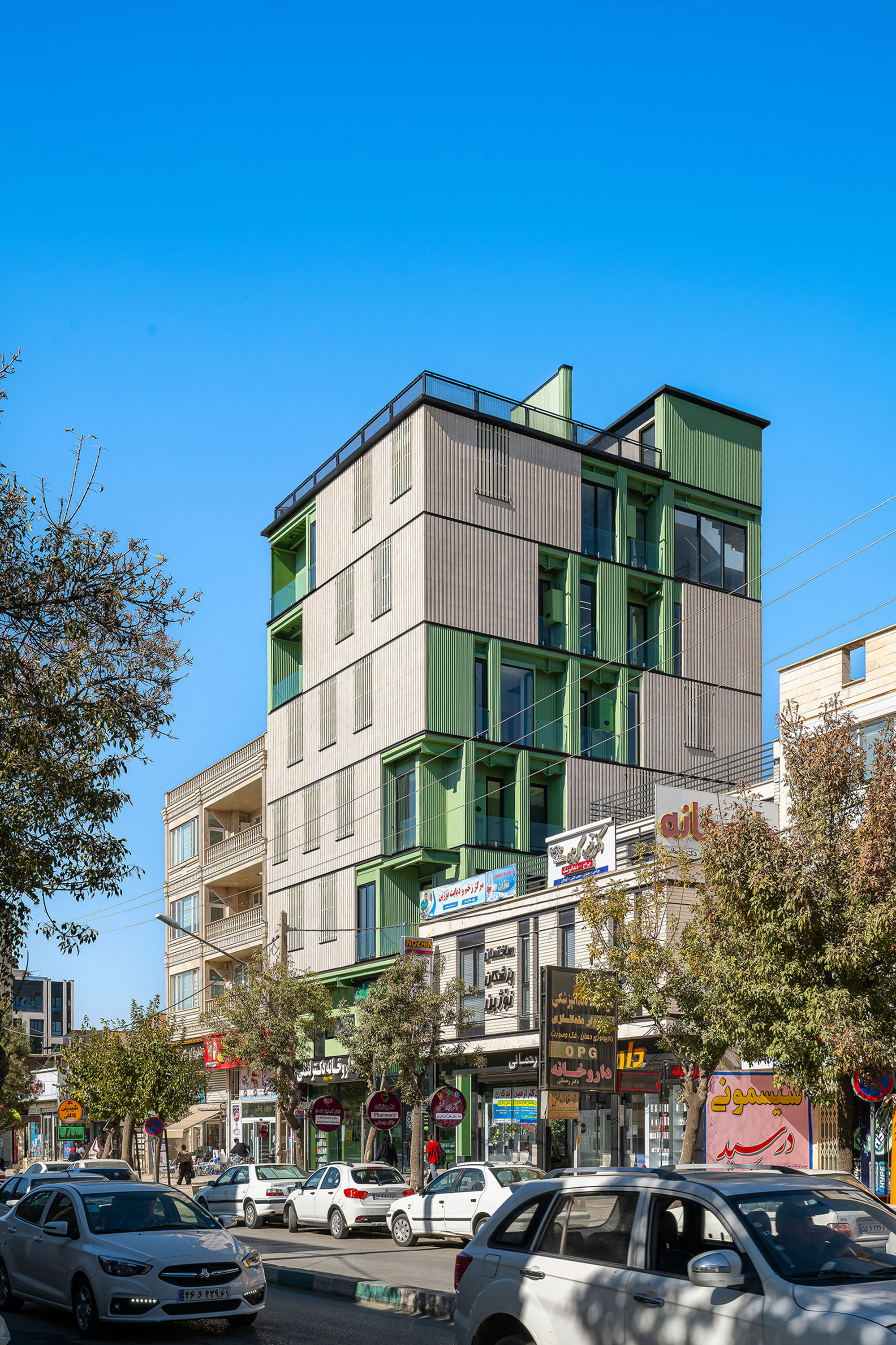 Street corner view of the stacked mixed-use block with green frames and grey cladding, parked cars and bare trees below a clear blue sky