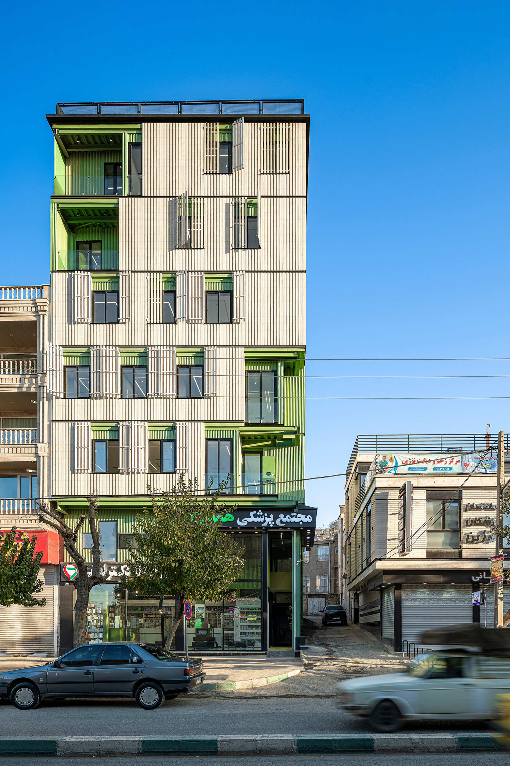 Front elevation of the tower with vertical ribbed cladding and green recesses, a tree and a passing car at street level