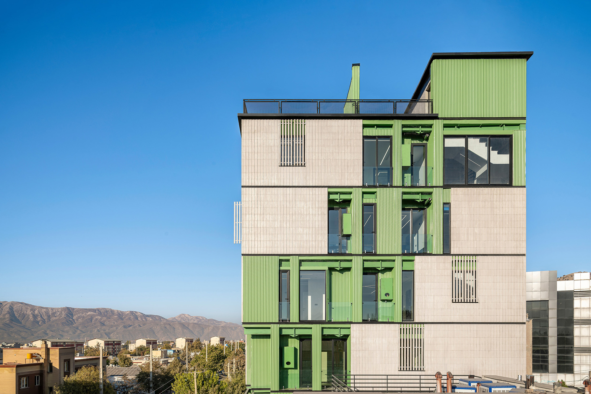 Side elevation of the stepped volumes with green panels and louvred openings against distant mountains in low afternoon sun
