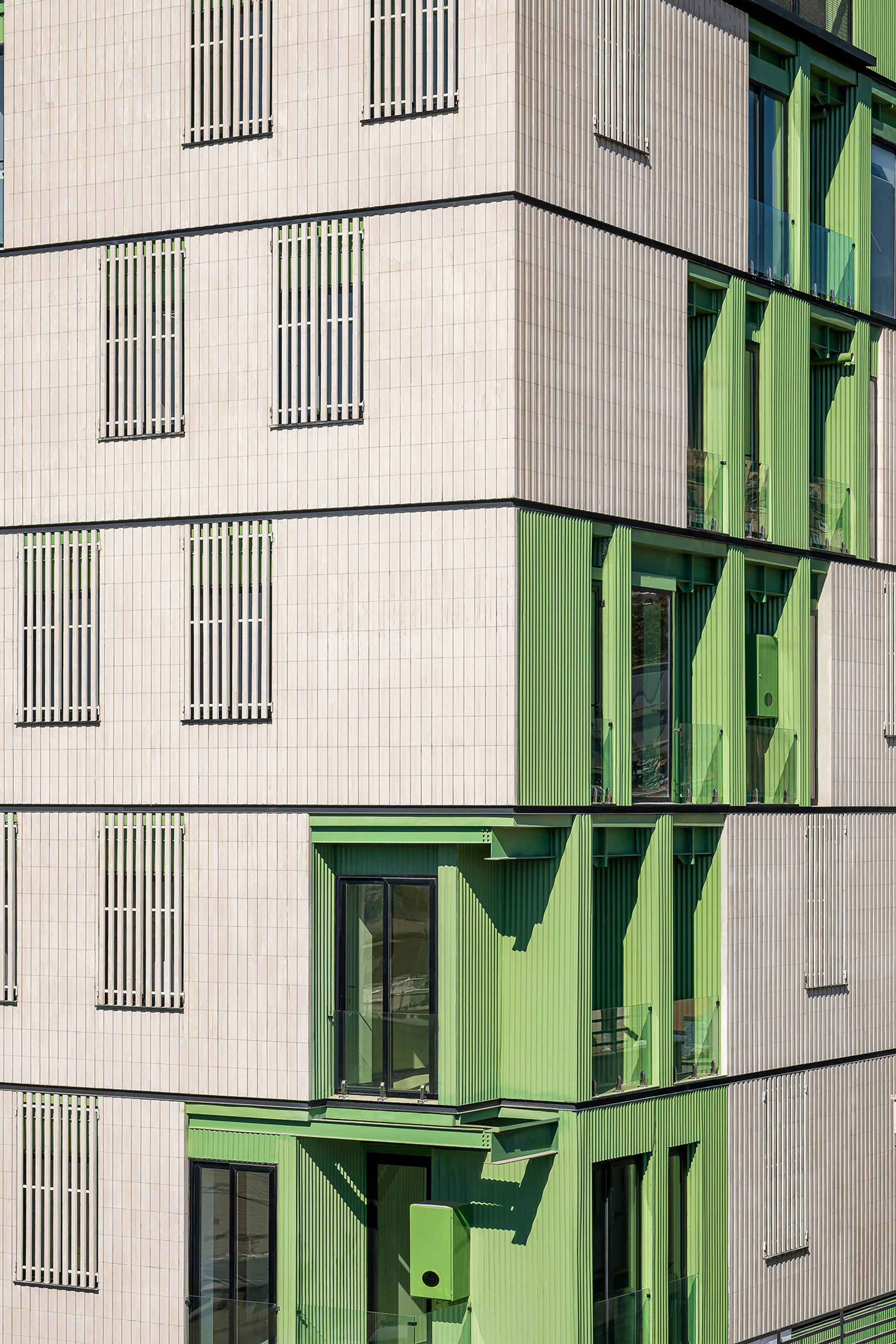 Close-up of the facade corner where green fluted panels and balconies interlock with pale cladding and slatted window screens in sunlight