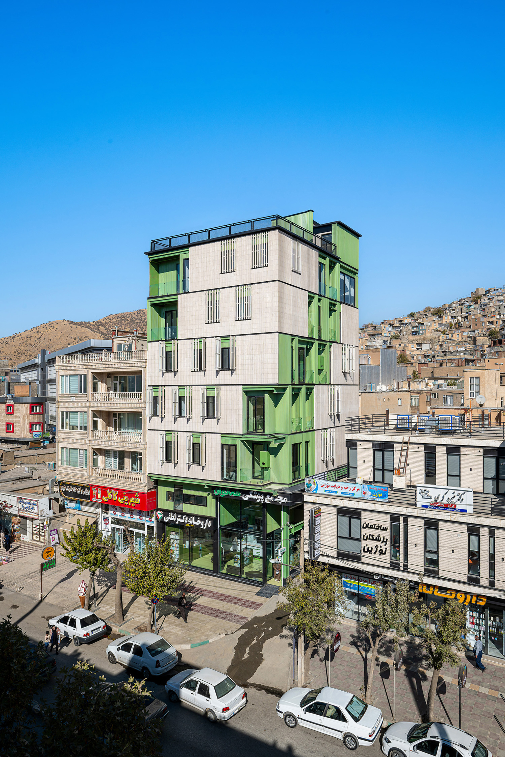 Elevated view of the green and grey tower over the street, with parked white cars, trees and hillside housing behind