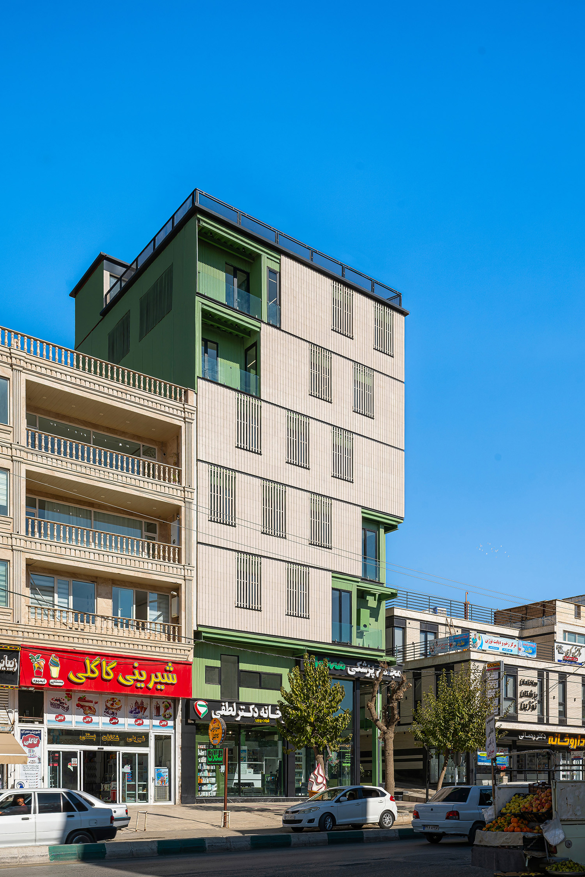 Street view of the five-storey facade with beige vertical cladding, slatted window screens and green upper terraces under a clear blue sky