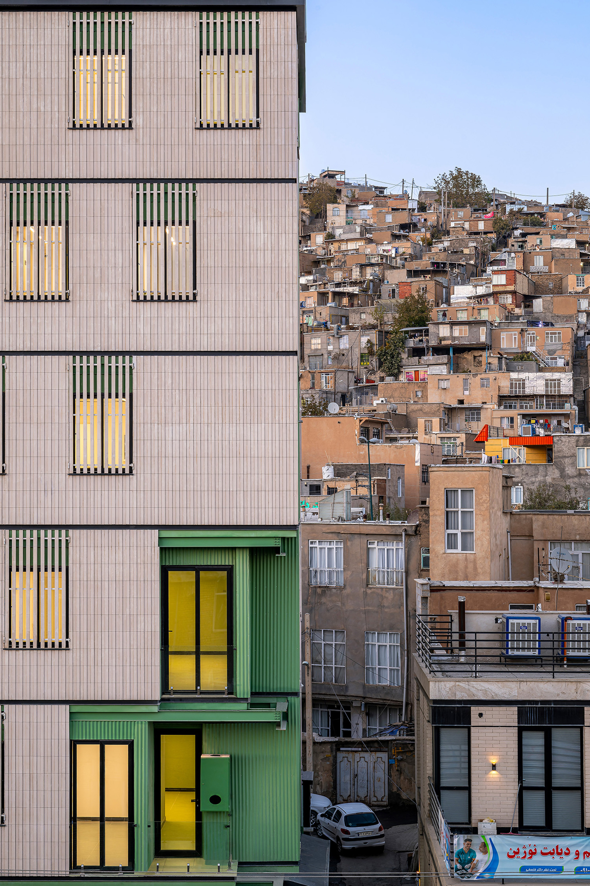 Side facade with yellow-lit windows and green corrugated panels facing a hillside of densely packed houses at dusk
