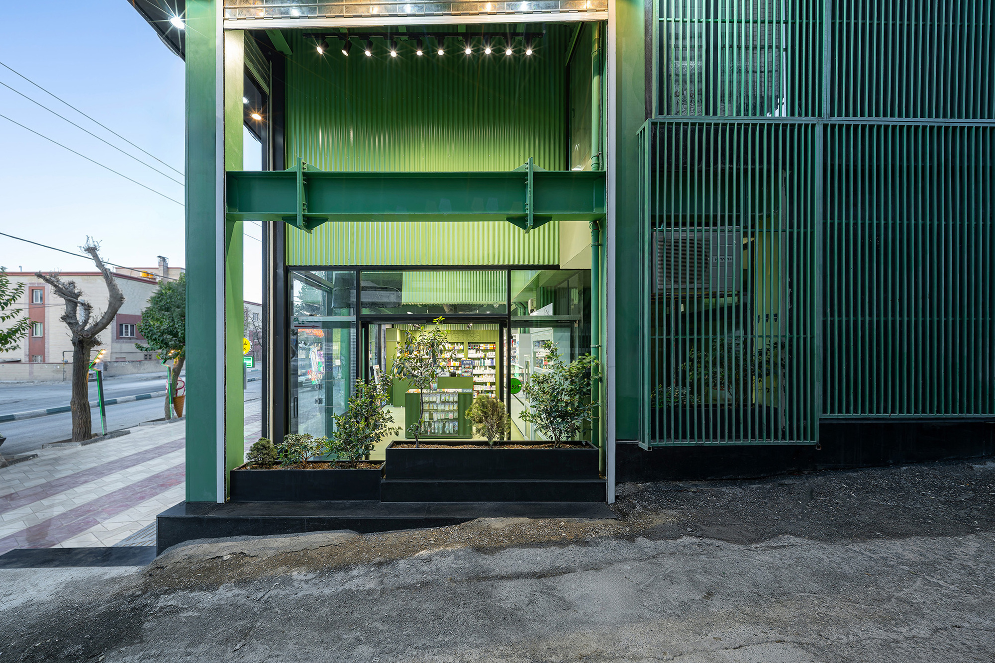 Ground-floor pharmacy entrance with green steel beam, planted black bed and slatted green screen at dusk
