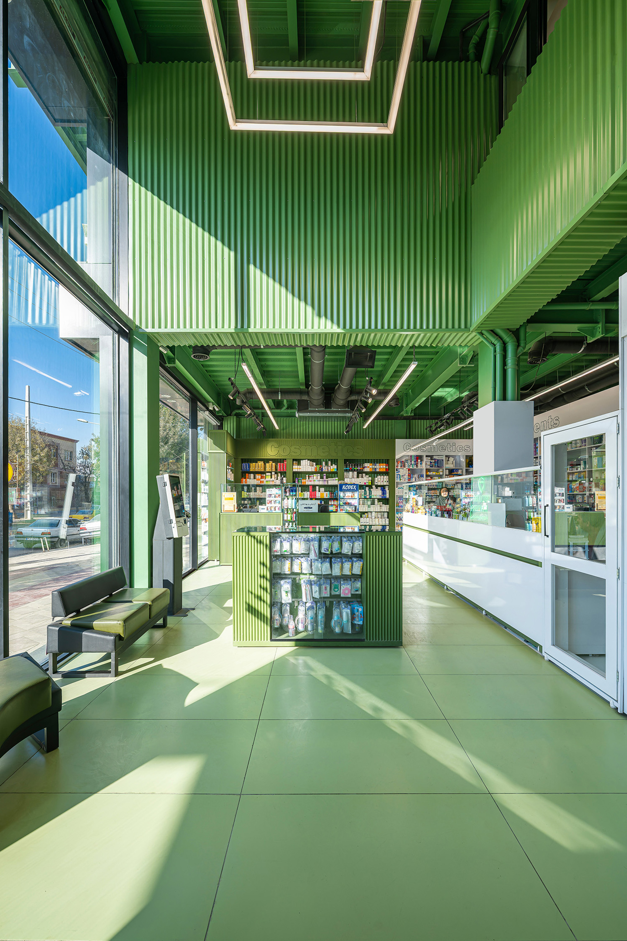 Pharmacy interior with green corrugated walls, green tile floor, a central display case and sunlight falling through the tall glazing