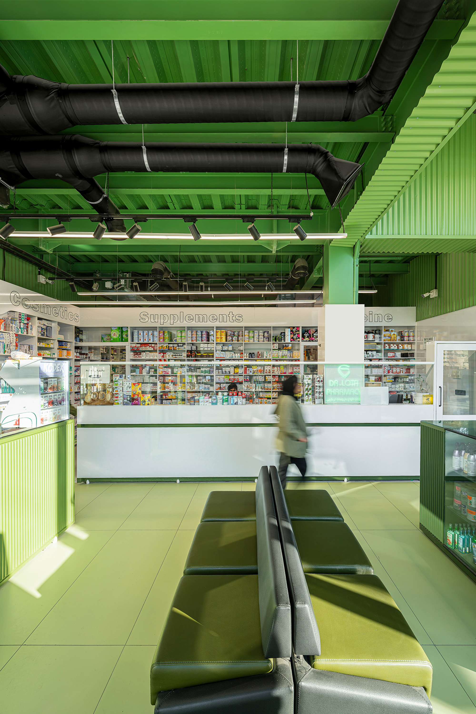 Pharmacy interior with black ductwork under a green ceiling, a white counter, shelves of supplements and a person walking past leather benches