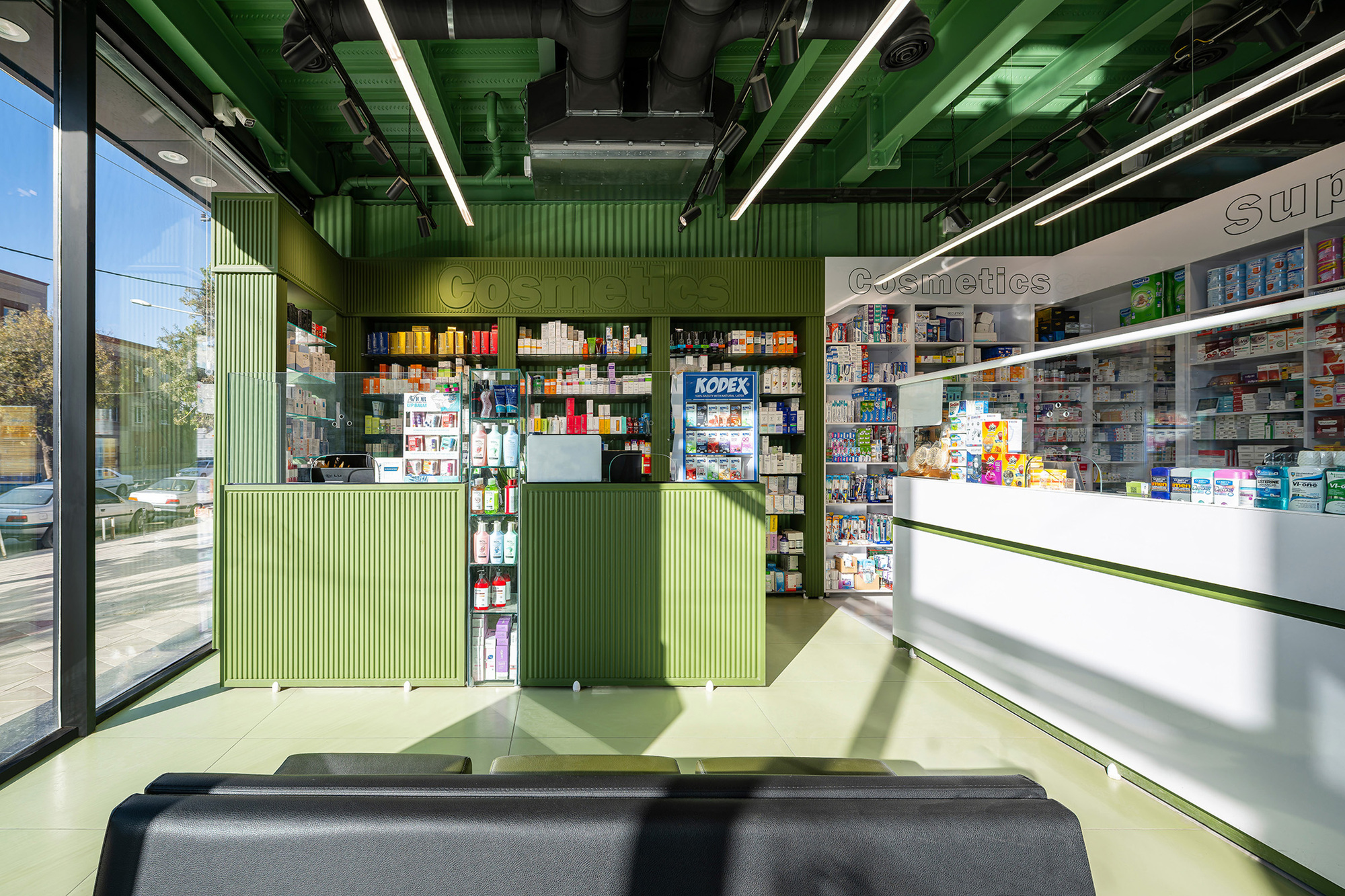 Pharmacy interior with green ribbed counters, shelves of cosmetics and a white sales desk under sunlit glazing and linear ceiling lights