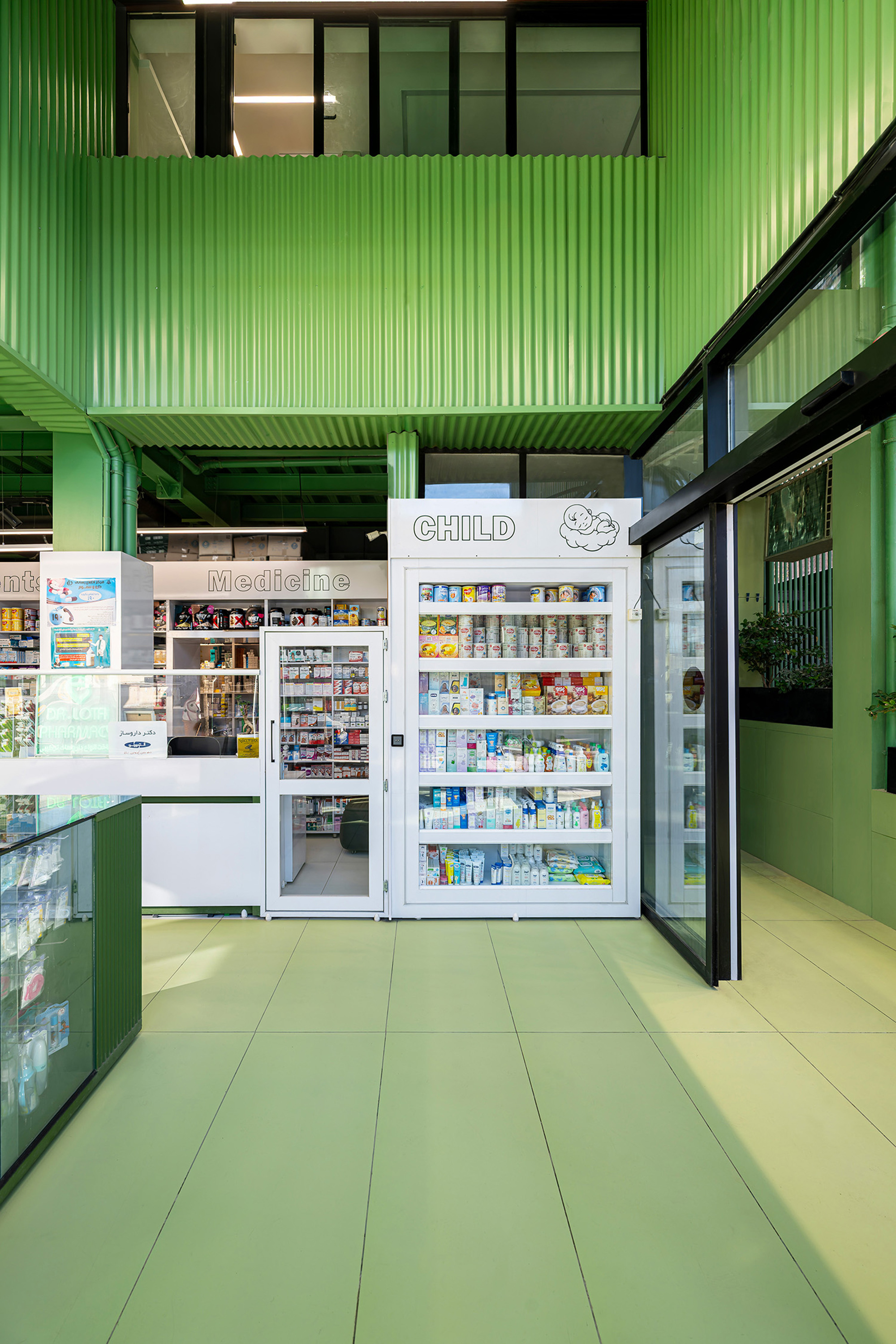 Green corrugated metal walls above a white child products cabinet and pale green floor tiles, with daylight from a glazed entrance