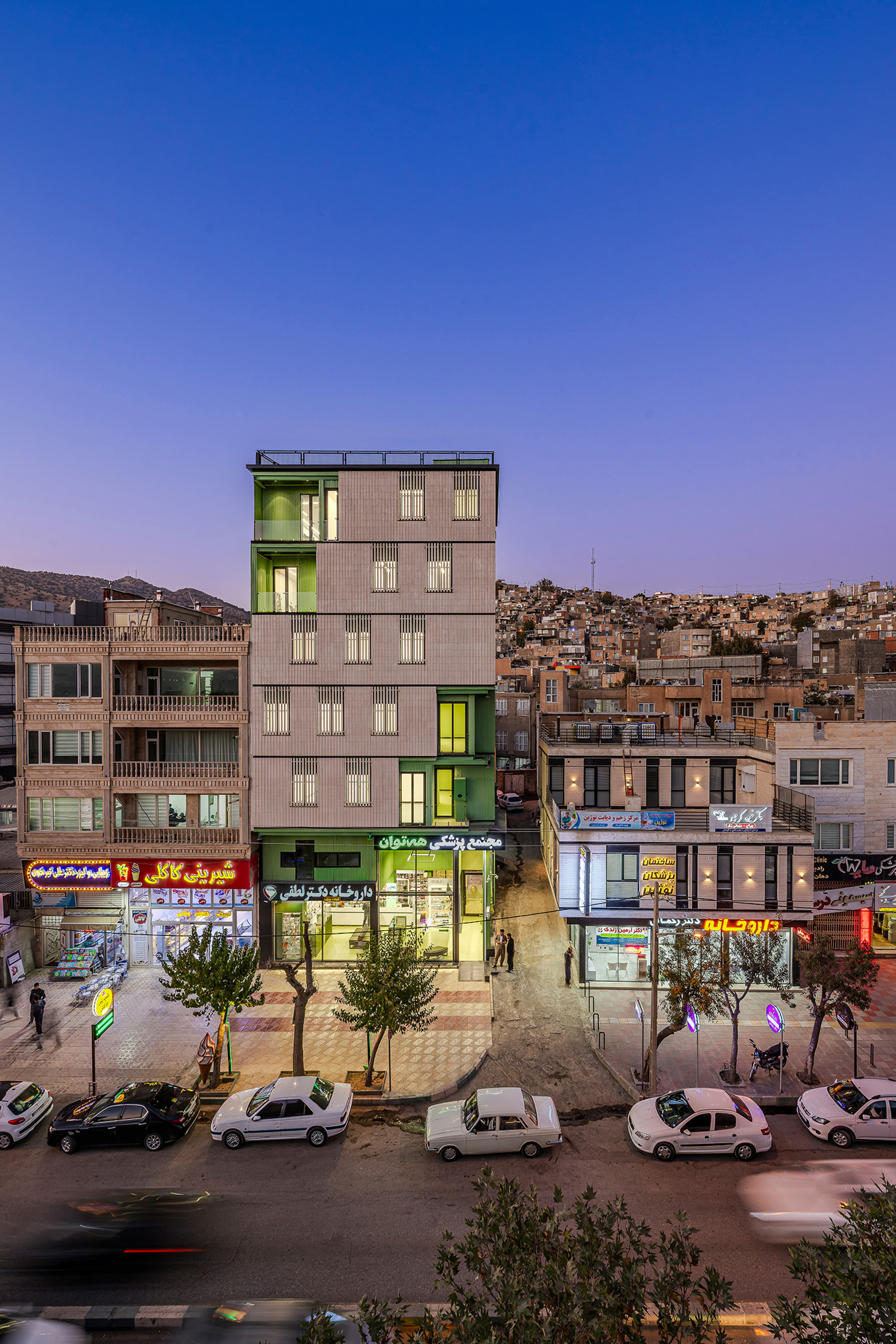 Street view of the narrow multi-storey block with green insets and a pharmacy at its base, glowing under a dusk sky