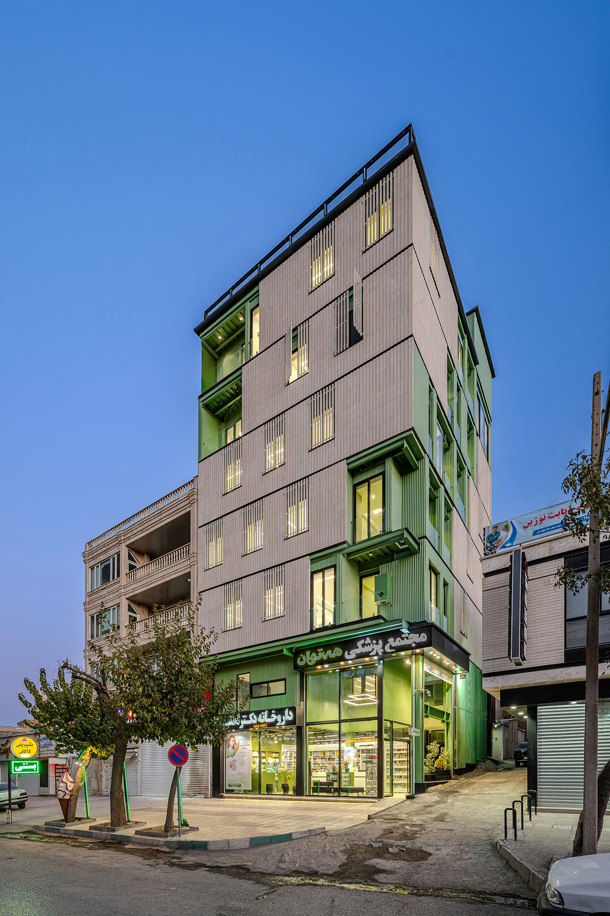 Corner view of the tall building with slatted facade panels and green frames, lit shopfront below and trees on the pavement at dusk