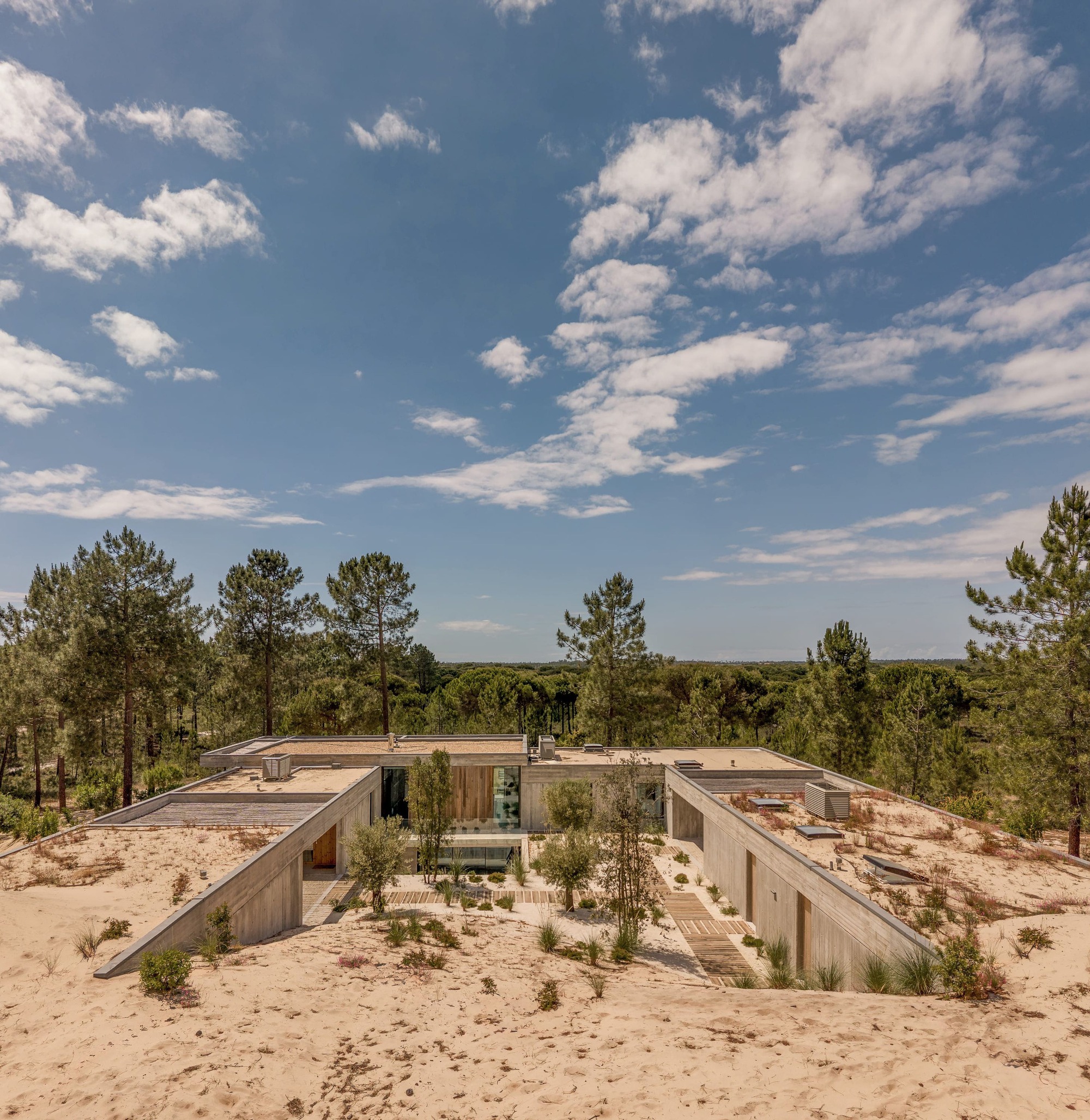 Aerial view of a flat-roofed board-formed concrete house sunk into sand dunes, with a planted courtyard under scattered clouds