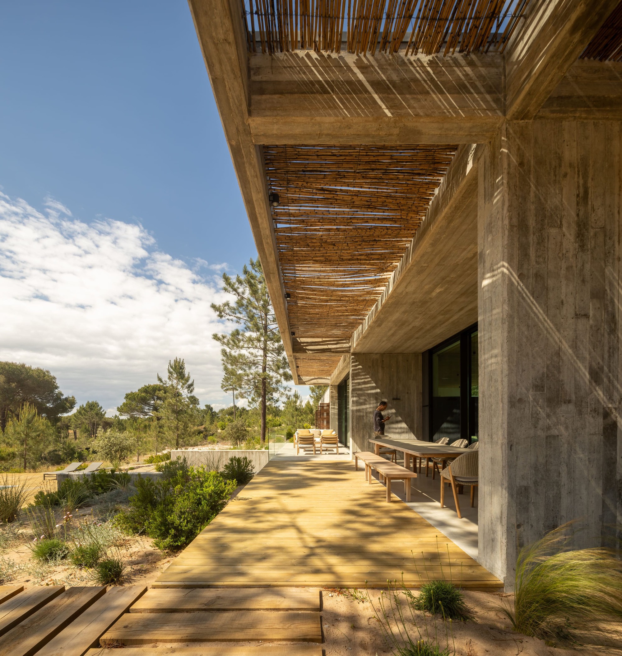 Covered terrace with board-marked concrete columns, a reed screen ceiling and a timber deck casting striped shadows in afternoon light