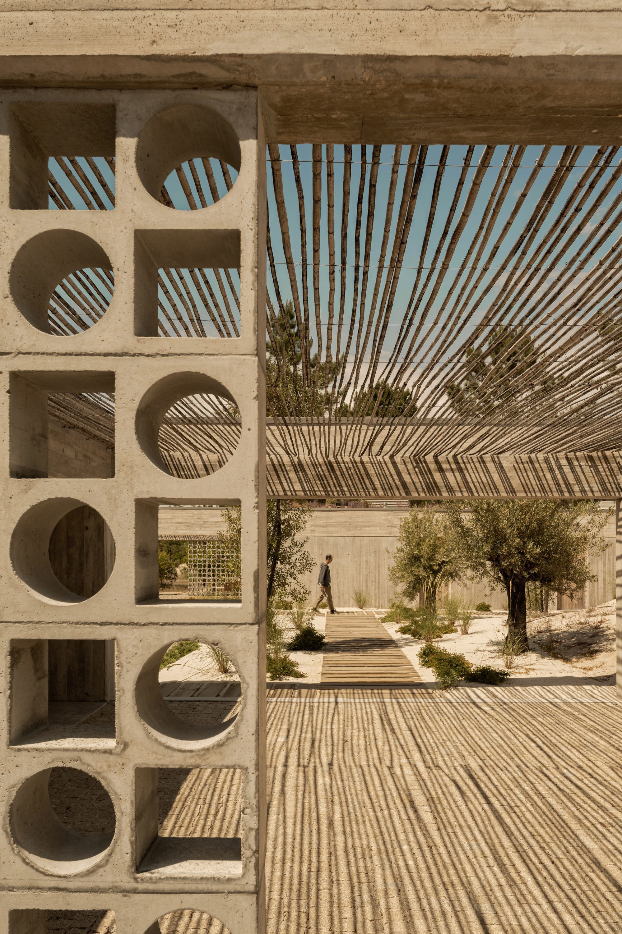 Perforated concrete block screen with round and square openings framing a reed pergola and a person walking a timber path