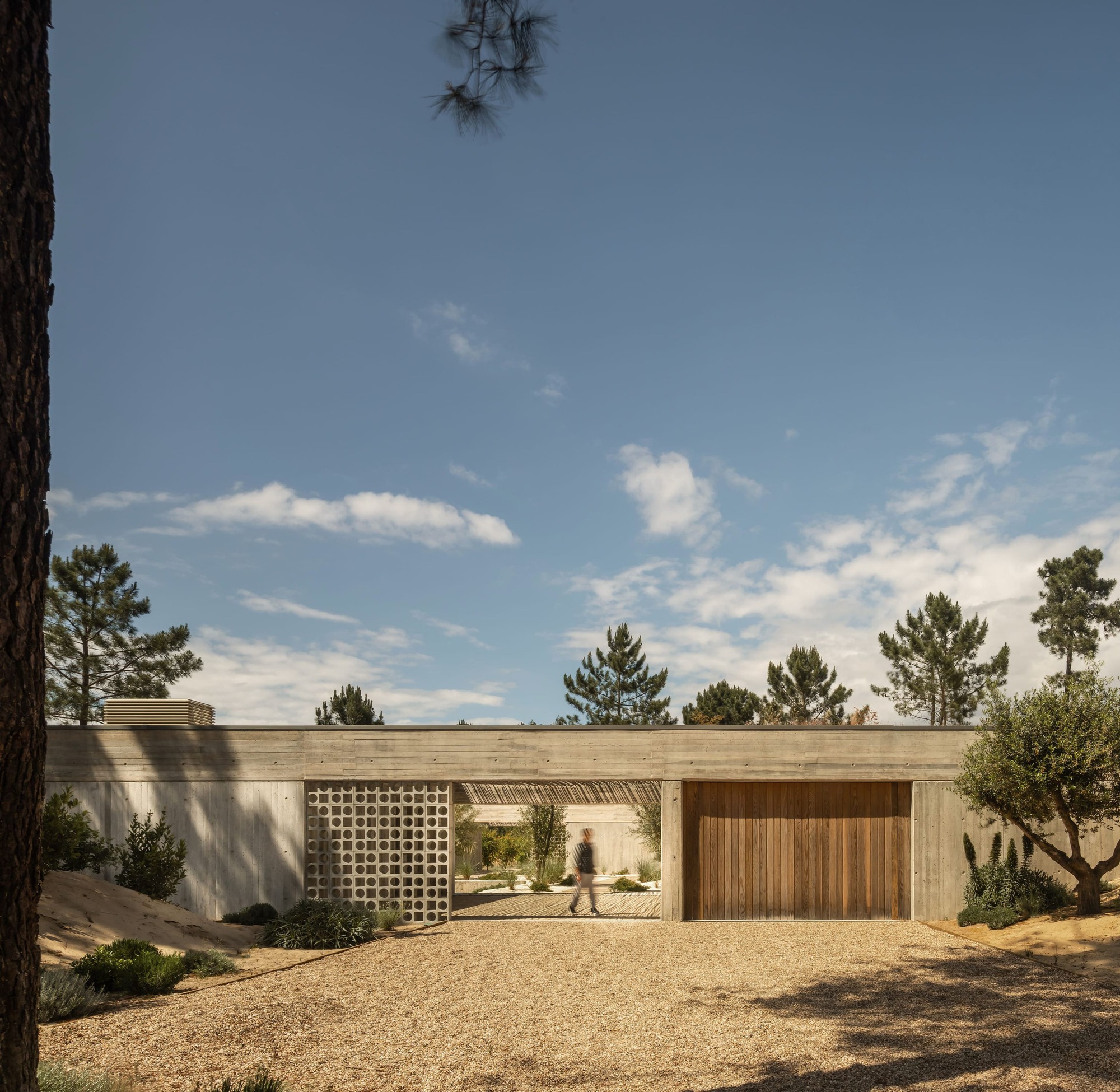 Street-side concrete wall with a patterned block screen and timber garage door, a blurred figure crossing the gravel entrance