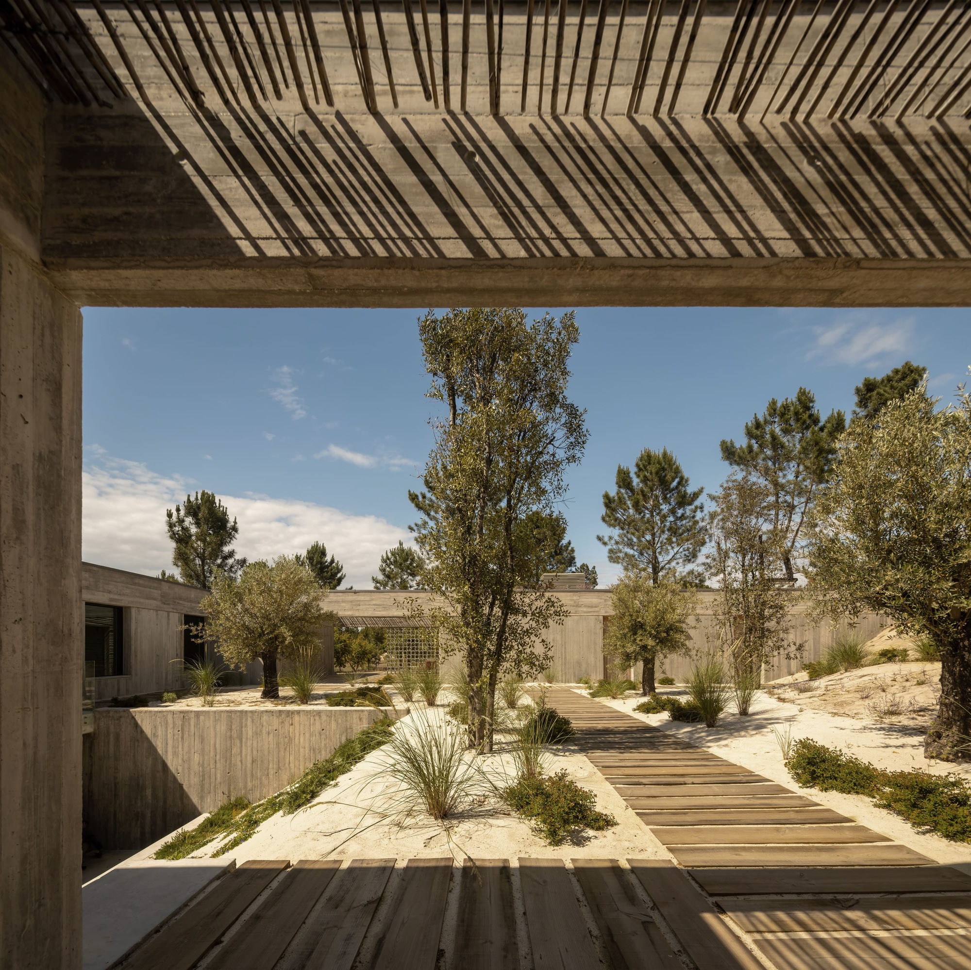 Looking through a concrete frame to a timber boardwalk crossing a sand courtyard, with slatted shadows cast across the ceiling