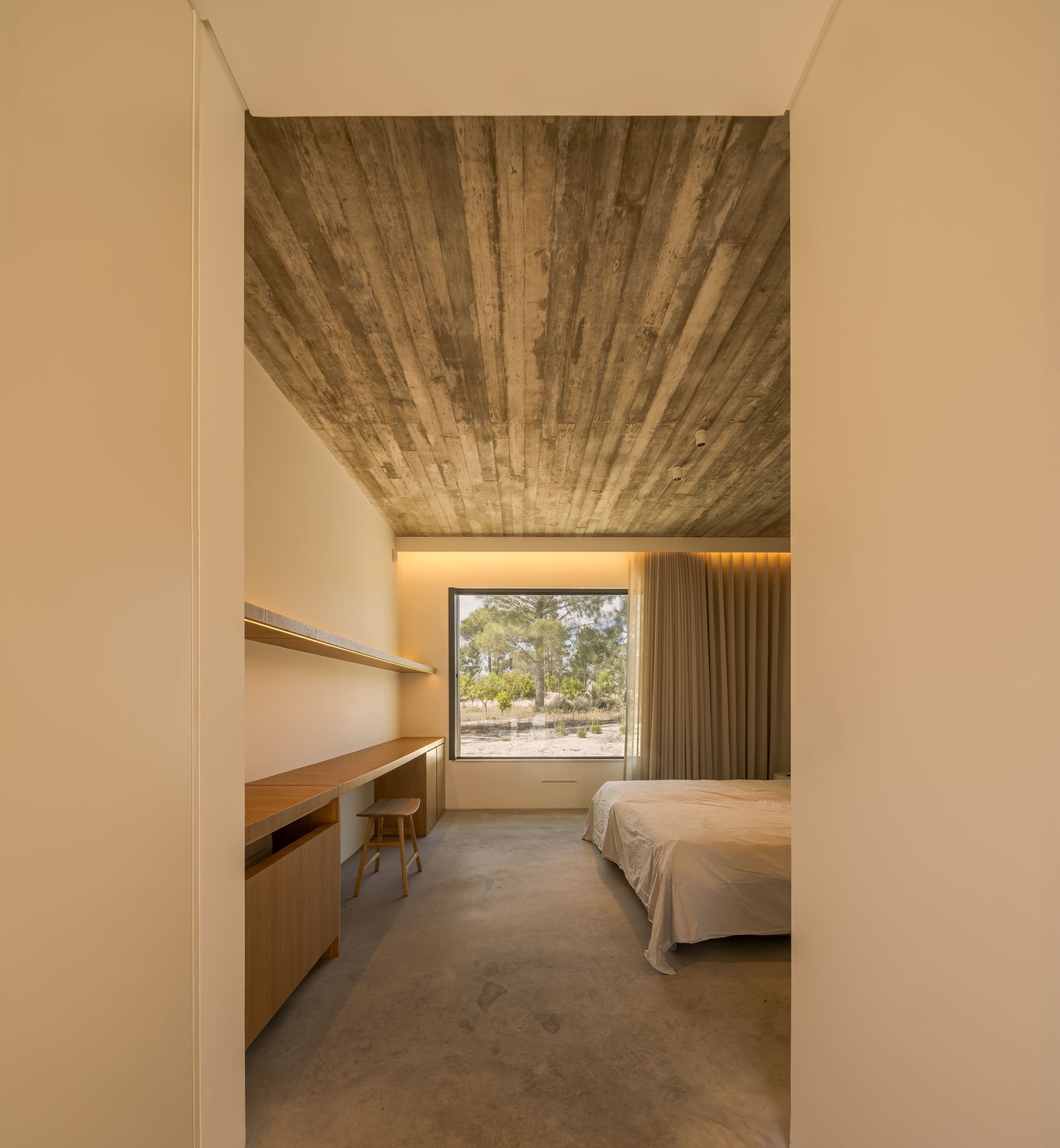 Bedroom with board-marked concrete ceiling, built-in oak desk and a square window onto pines, lit by a warm cove light