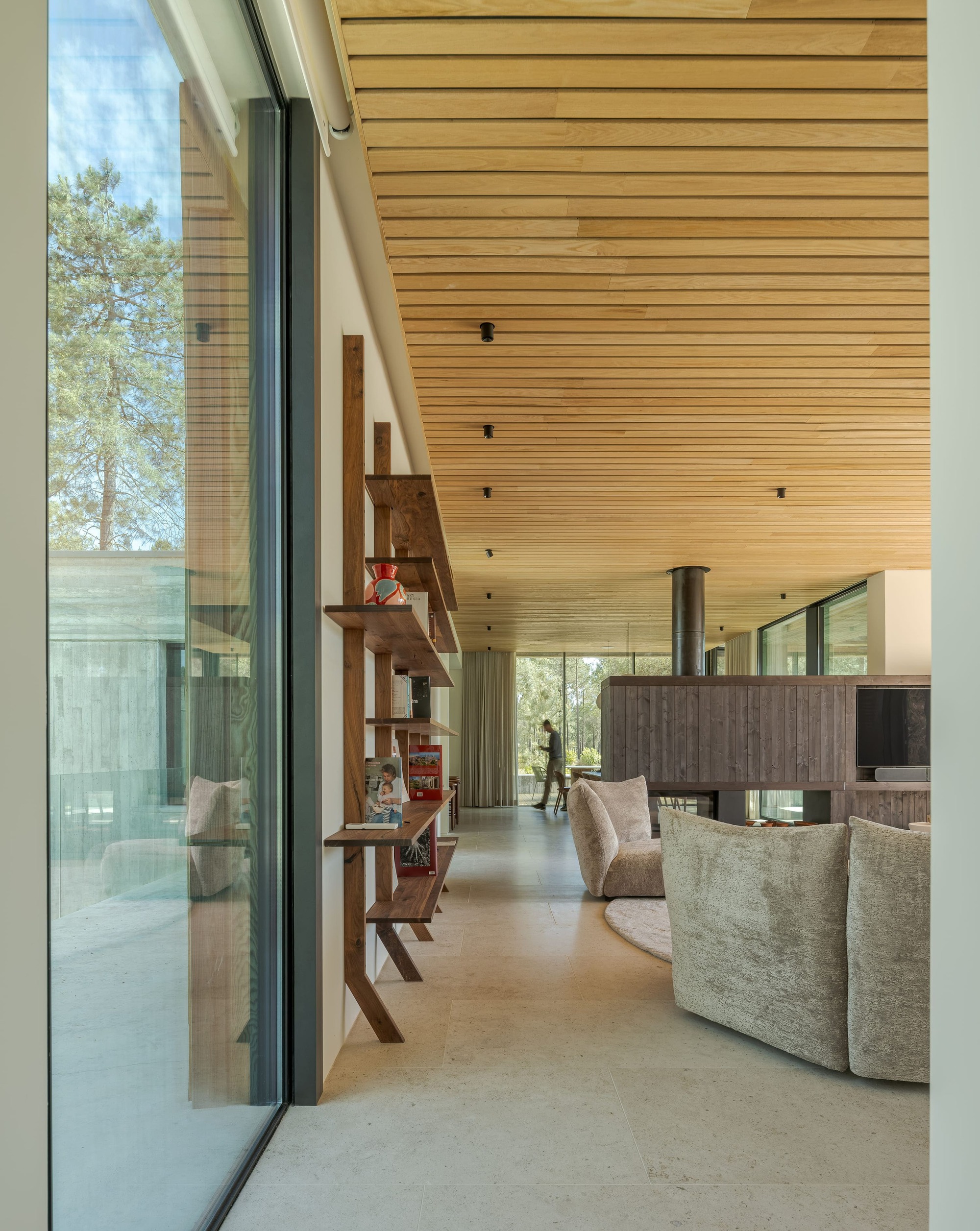 Living space with oak slat ceiling, walnut shelving beside floor-to-ceiling glass and a central steel flue, with a person walking beyond