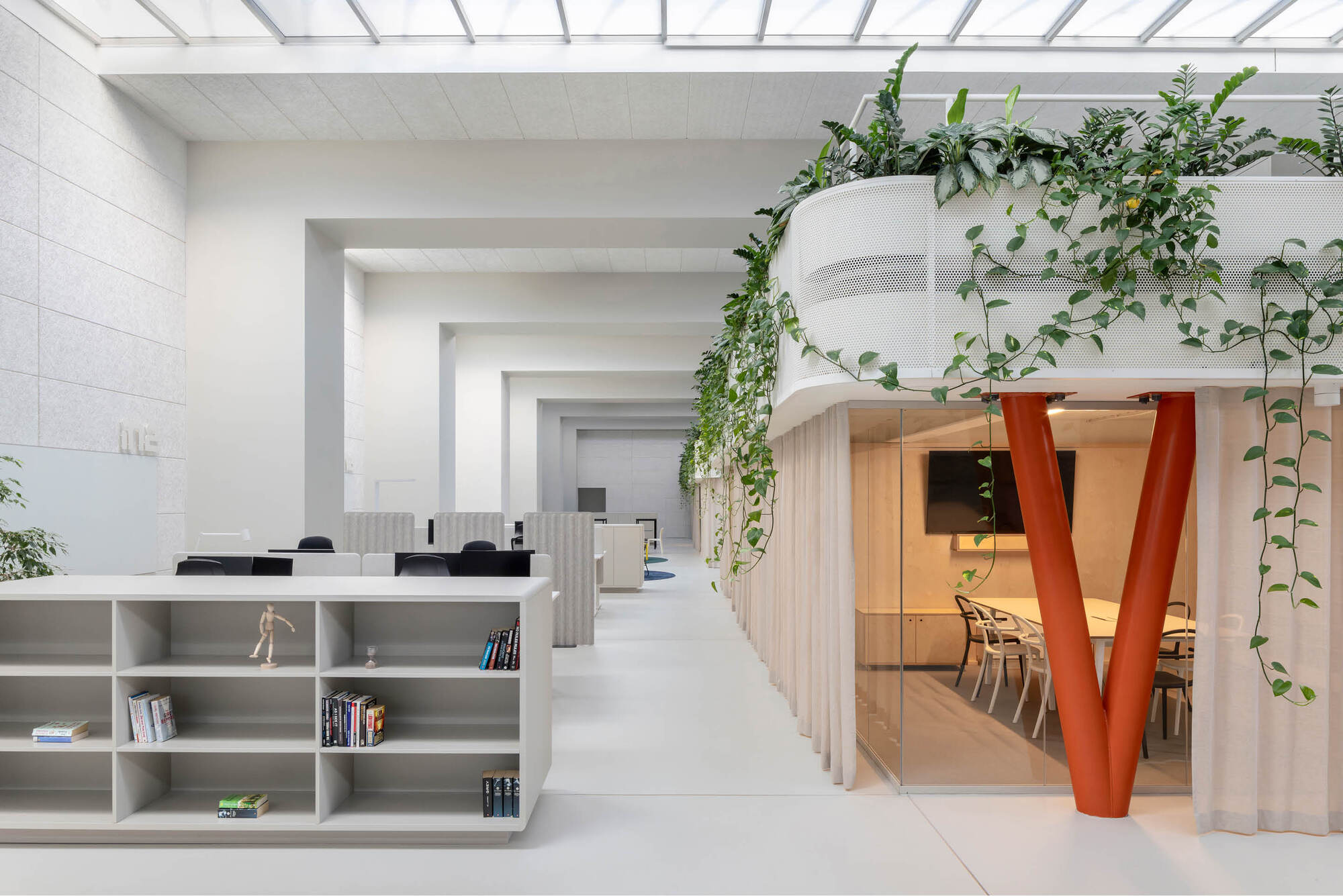 Open office aisle with a grey shelving unit and a glass meeting room under an orange V-shaped column draped in trailing vines