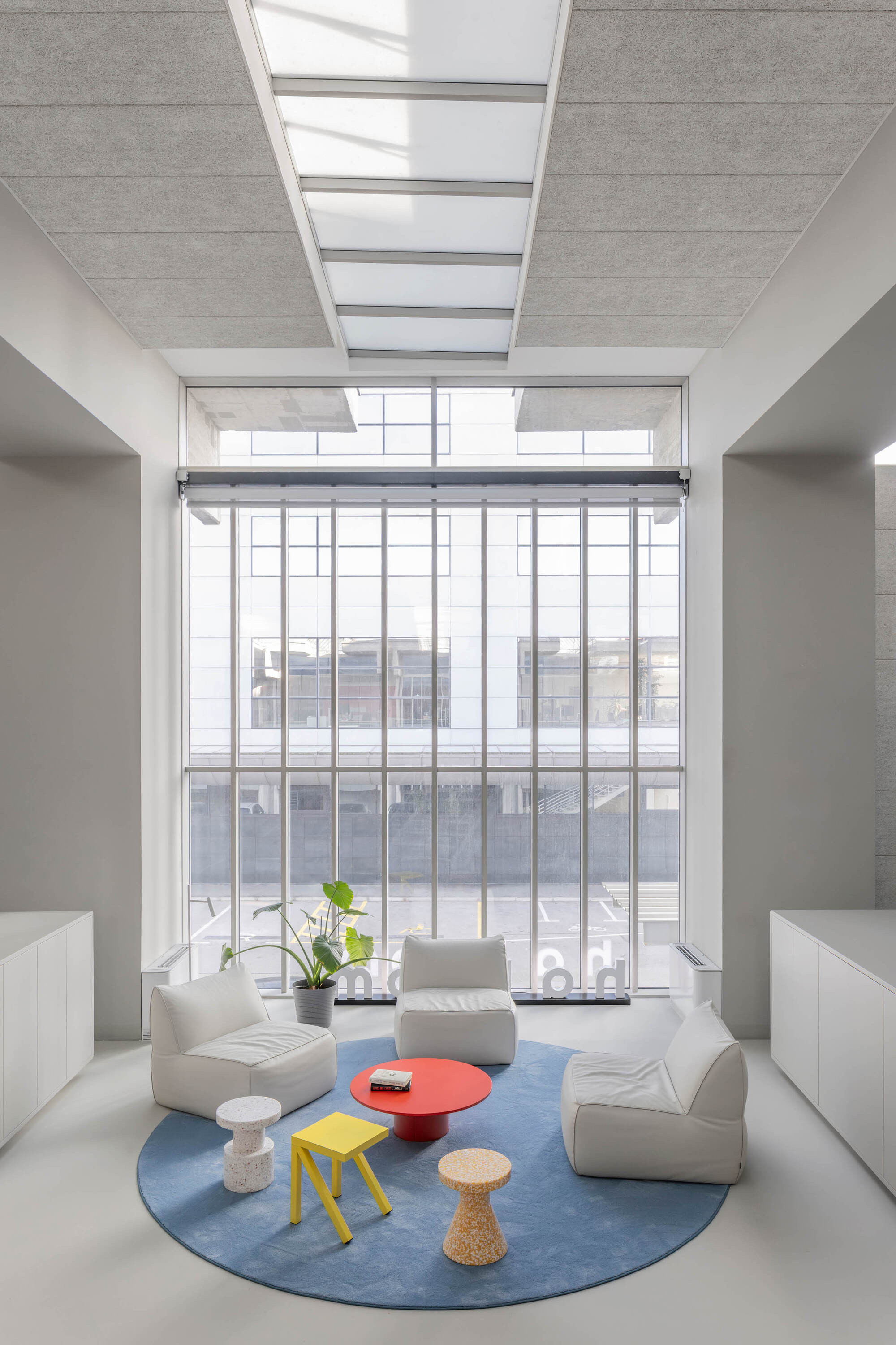 Double-height lounge with white armchairs and a red table on a round blue rug facing a tall gridded window