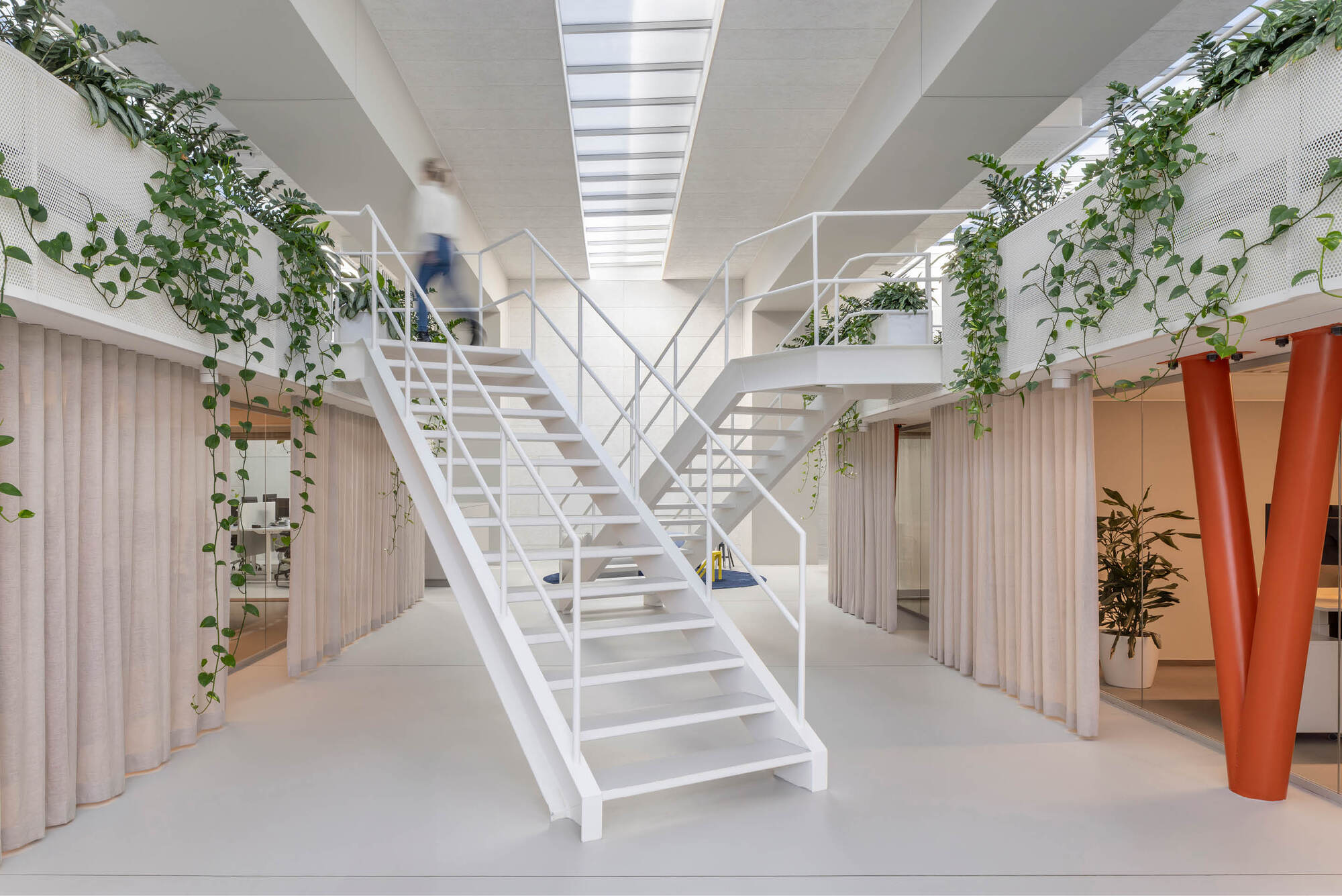 White steel staircase rising between planted balconies and pleated curtains, with a blurred figure climbing in skylight